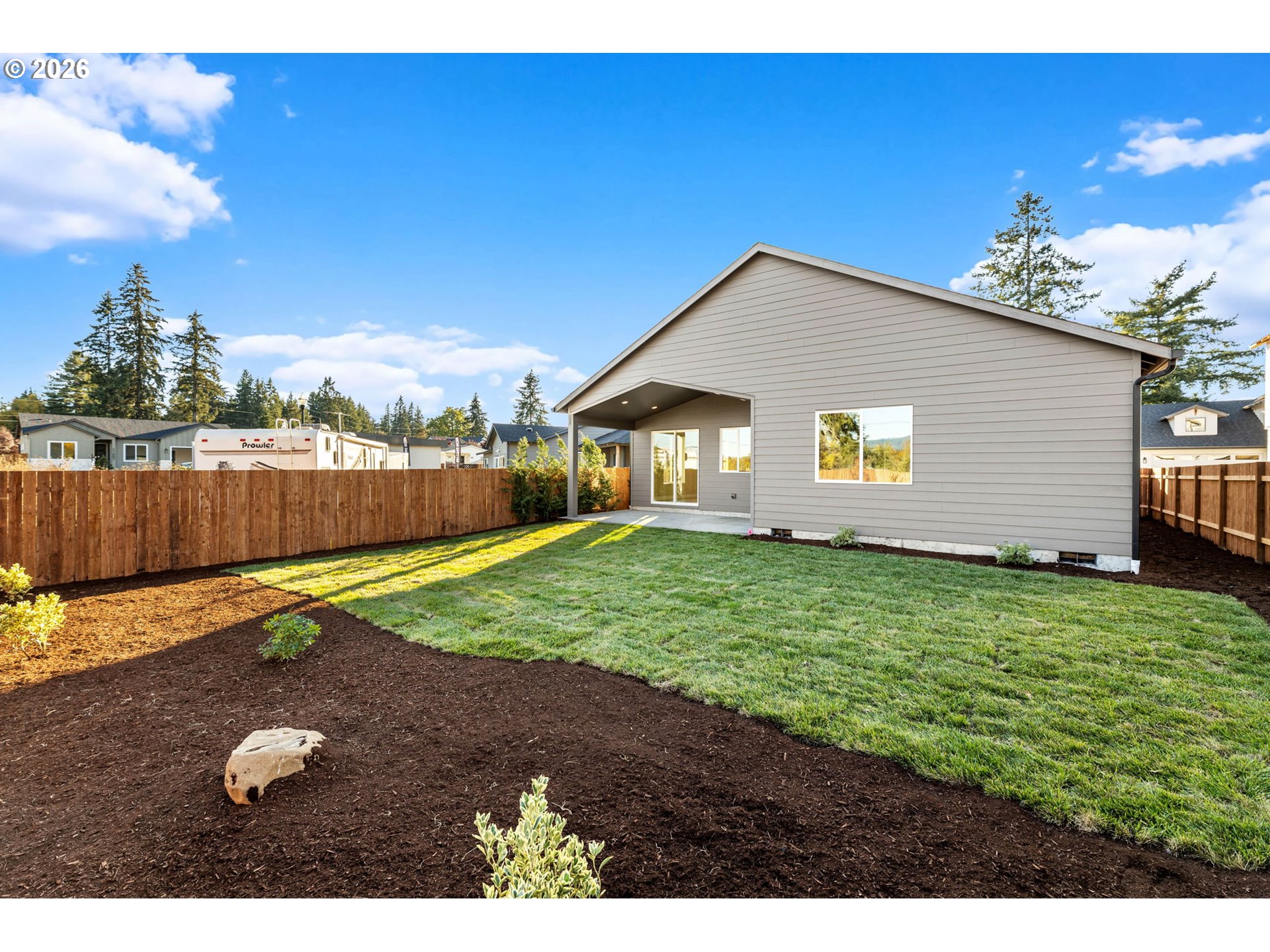 Rear of a gray-sided house with a covered patio, fenced lawn, and landscaped garden beds.