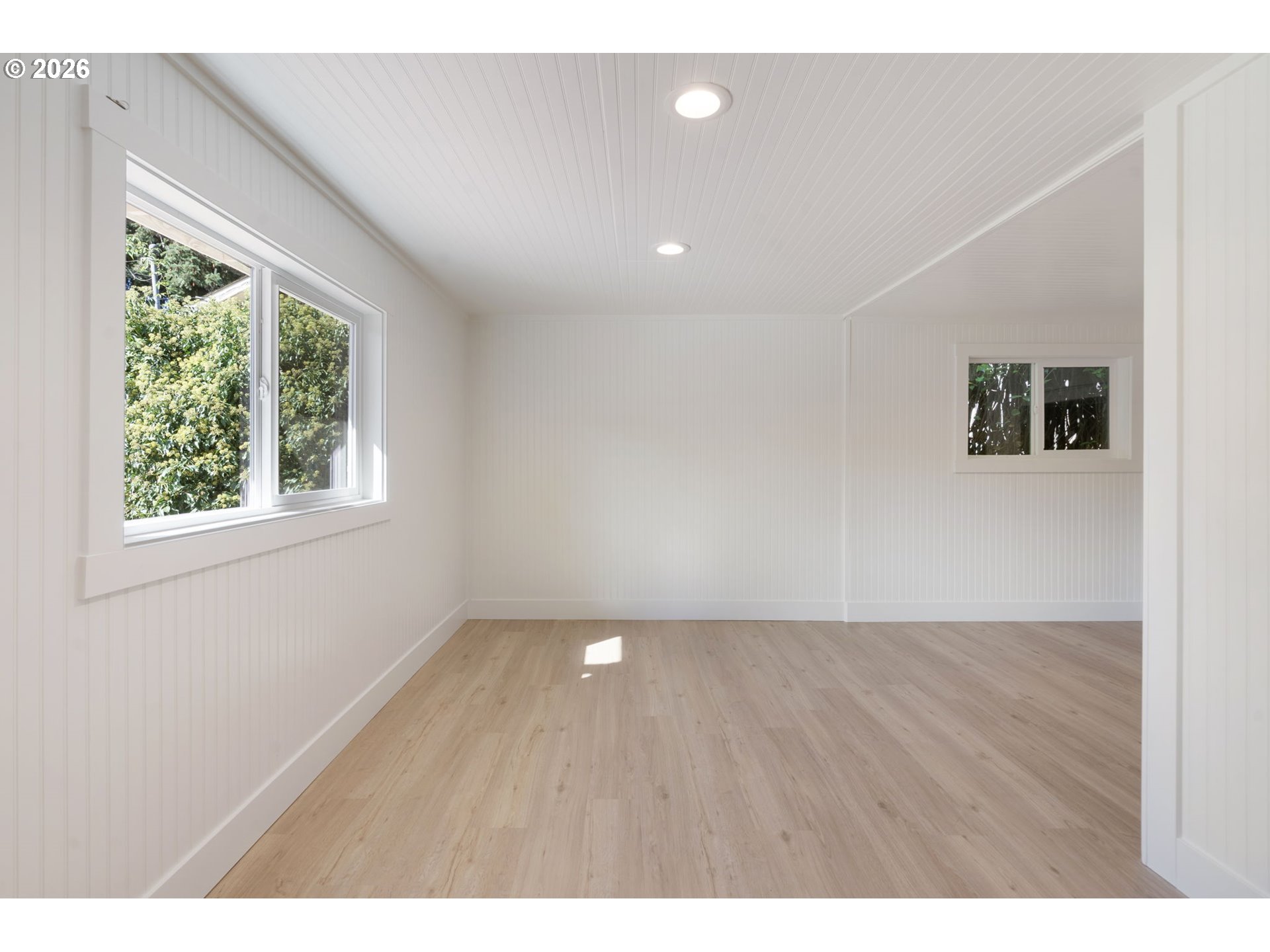 Empty bright room with white beadboard walls, light wood floors, and large windows.