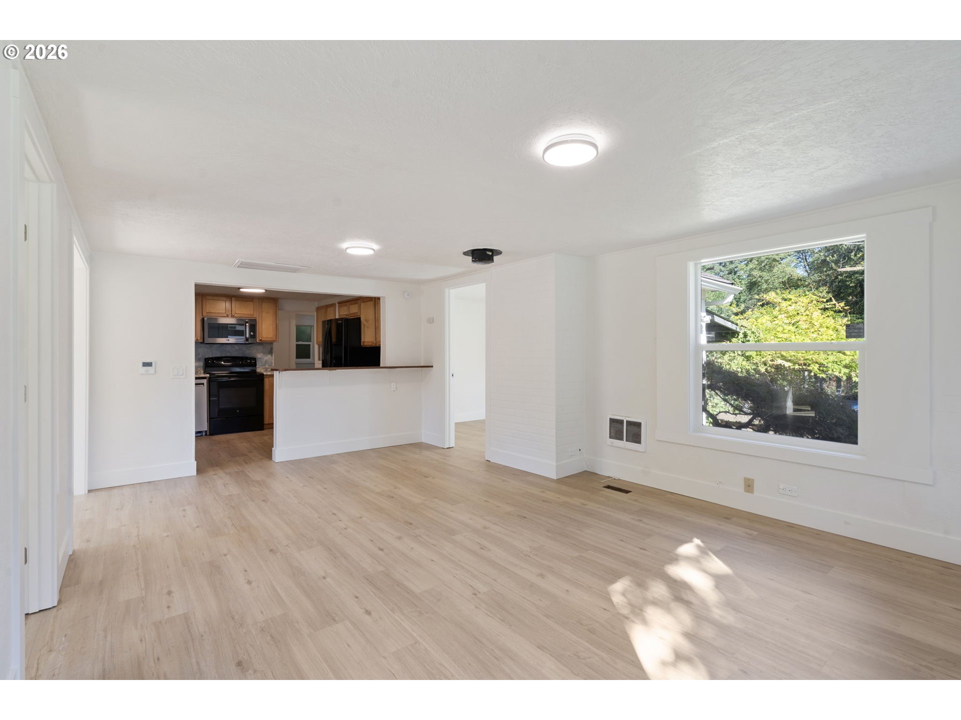 Unfurnished living room with light wood floors, a large tree-view window, and an open kitchen.