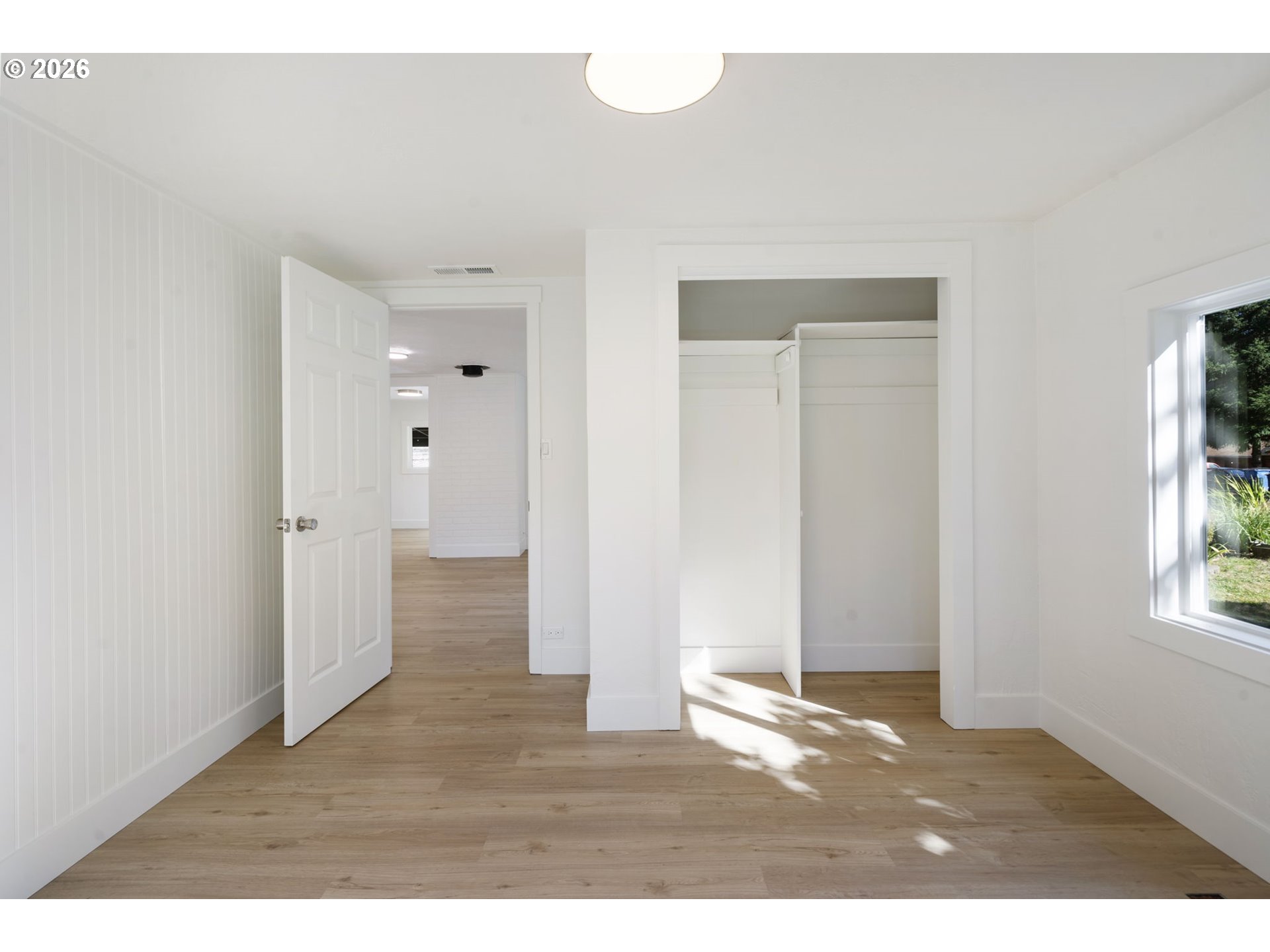 Vacant bedroom with an open closet, light wood floors, and sunlight falling through a window.