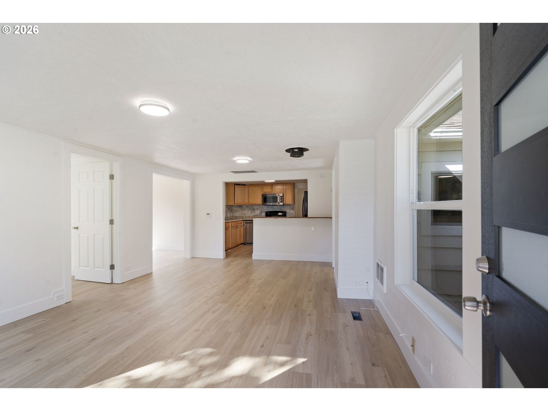 Empty living area with light flooring, a large window, and an open kitchen with wood cabinets.