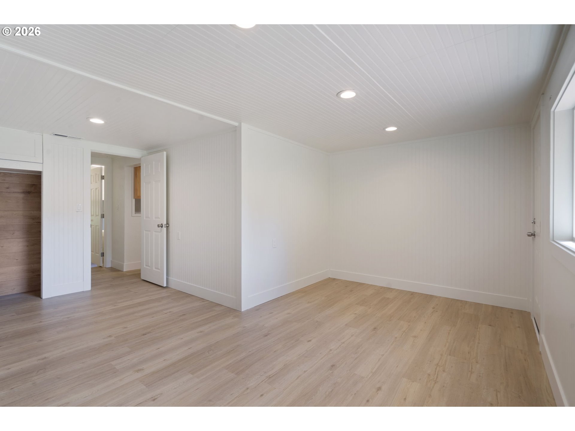 Empty room with light wood flooring, white paneled walls, recessed lights, and a large window.