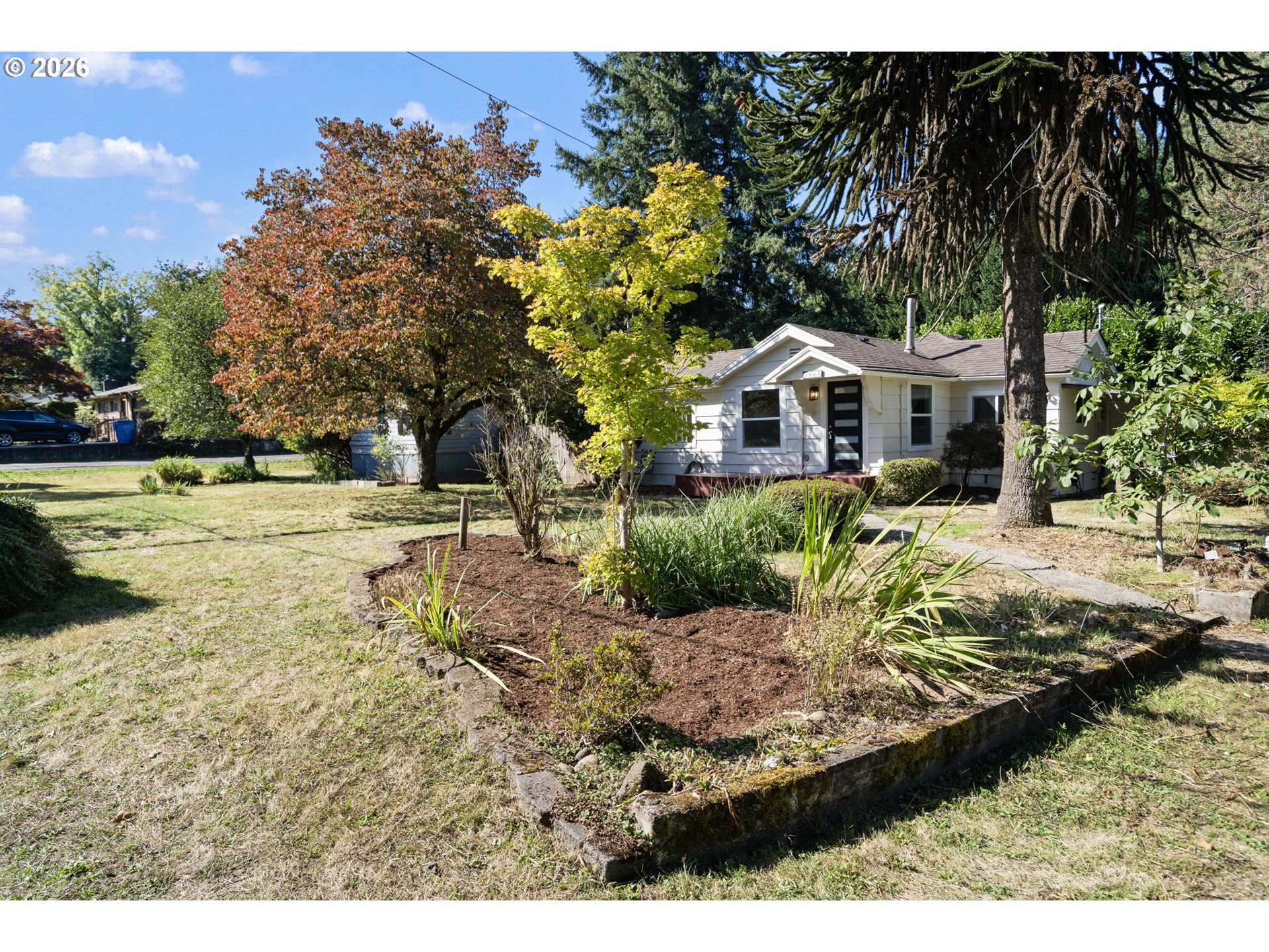 White single-story house behind a raised garden bed and mature trees.