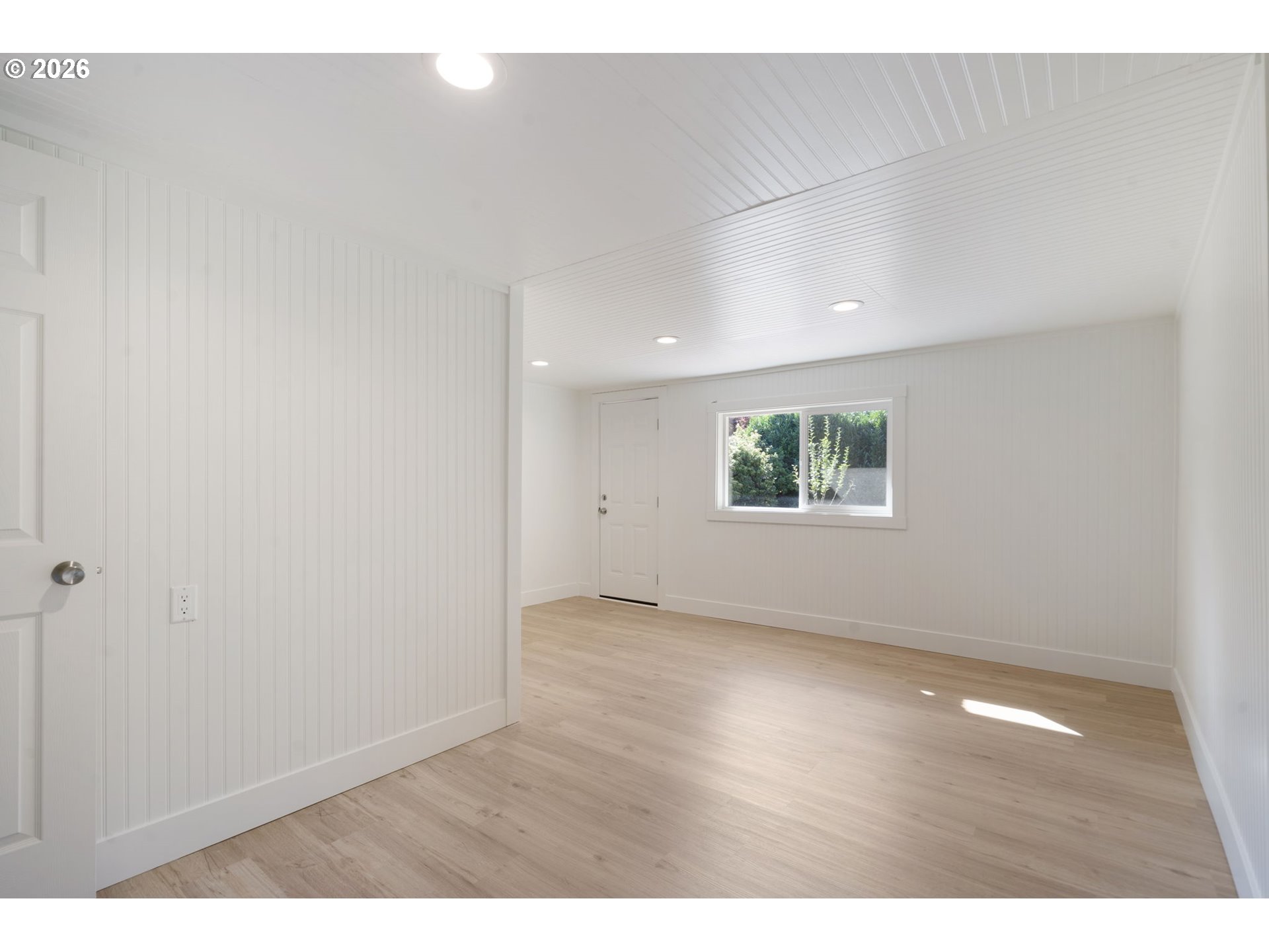 Empty room with white beadboard walls, light wood flooring, and a window overlooking greenery.