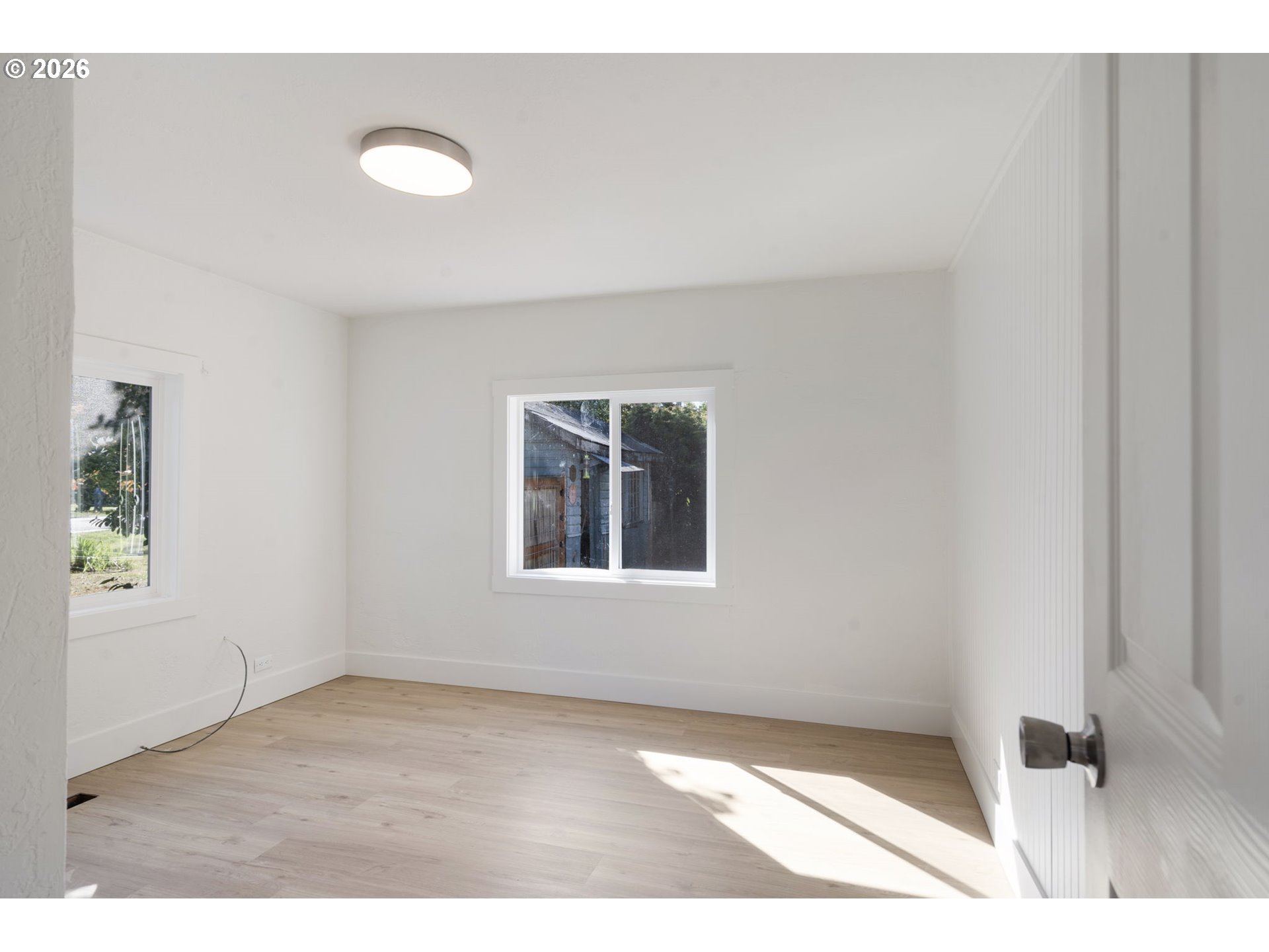 Empty room with pale wood floors, white walls, two windows, and sunlight across the floor.