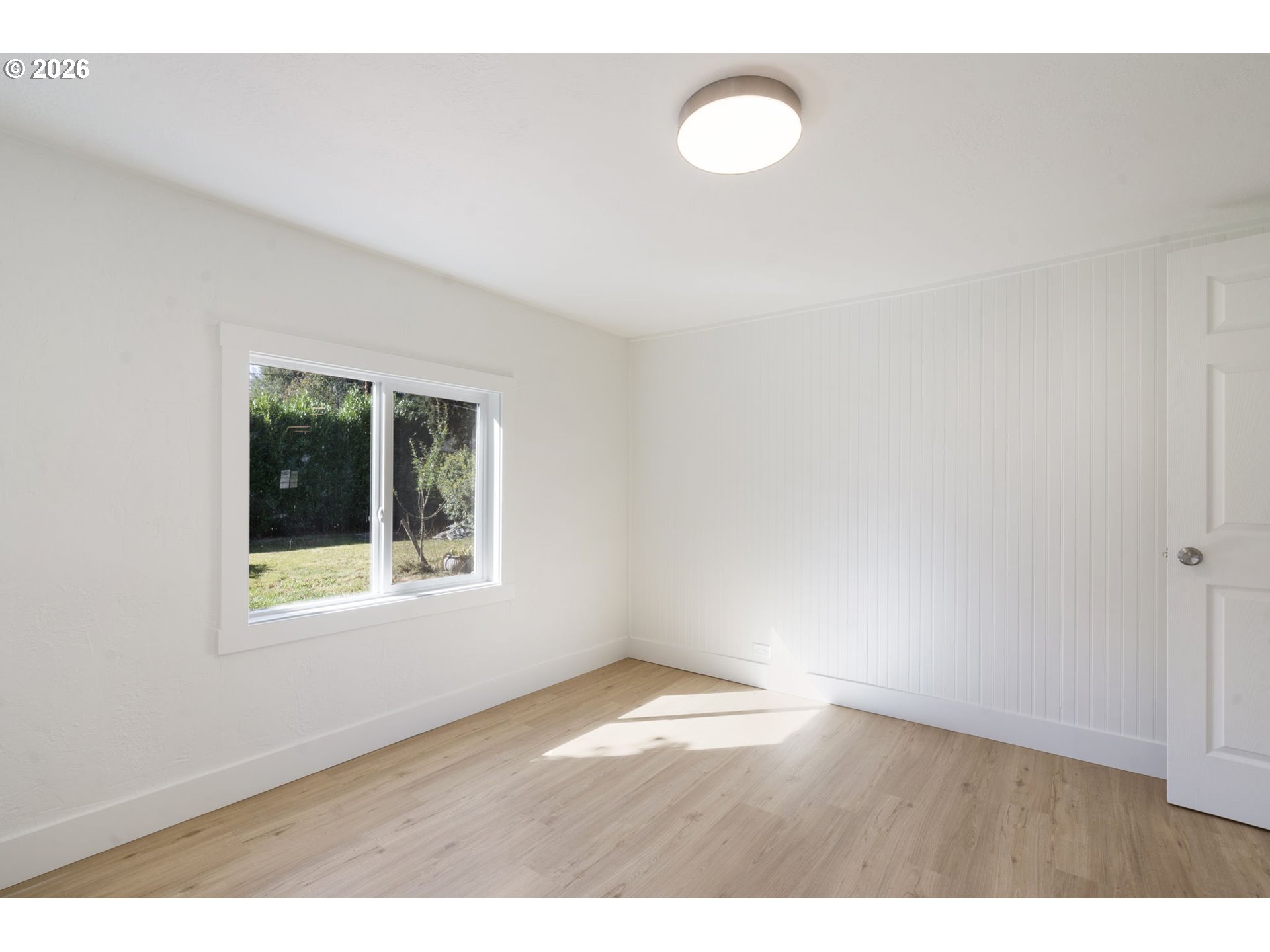Empty room with light wood floors, white beadboard wall, and a window overlooking the yard.