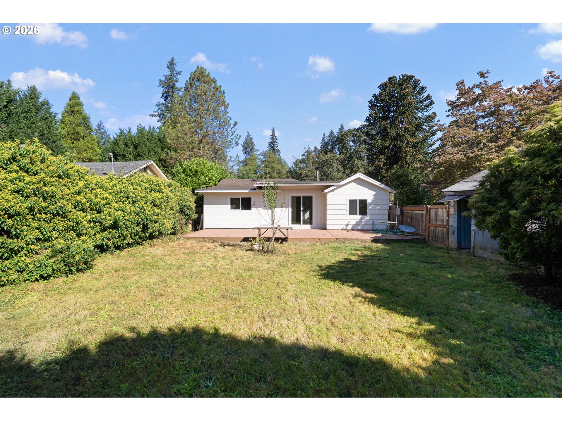 White single-story house beyond a grassy backyard, with a brick patio and tall hedges.