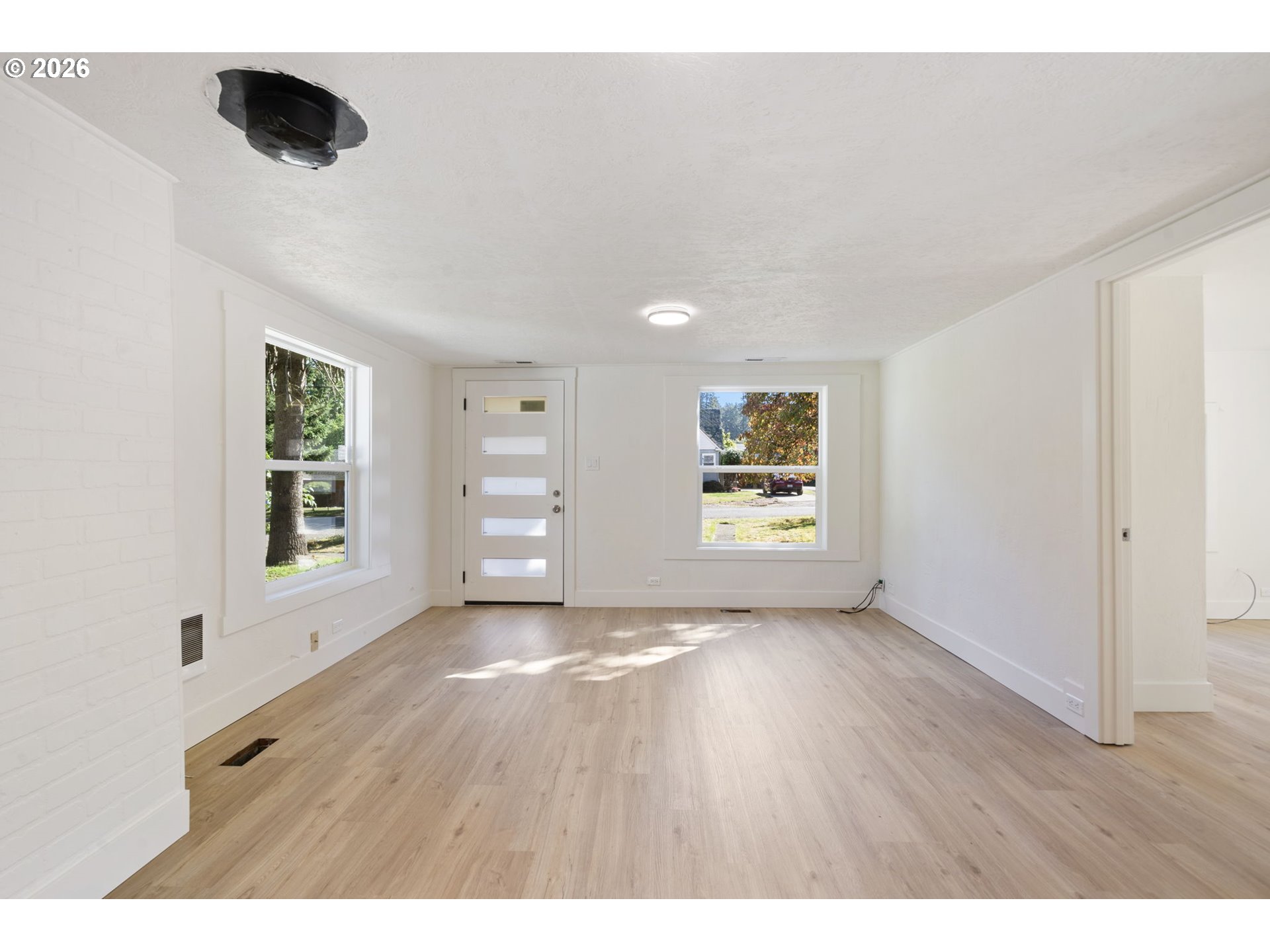 Empty living room with pale wood floors, two windows, and a front door with glass panels.