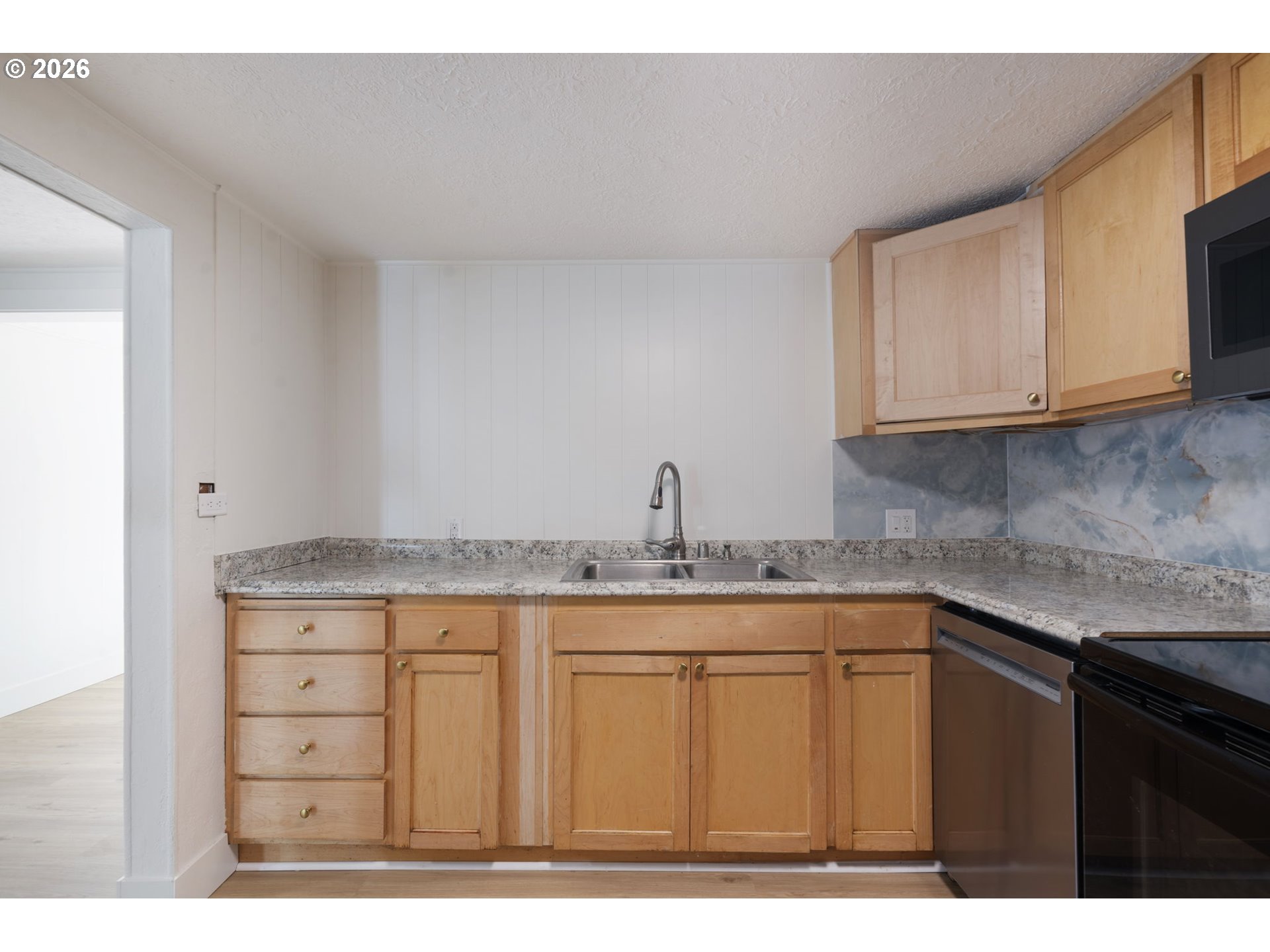 Kitchen with light wood cabinets, speckled counters, a double sink, and blue-gray backsplash.