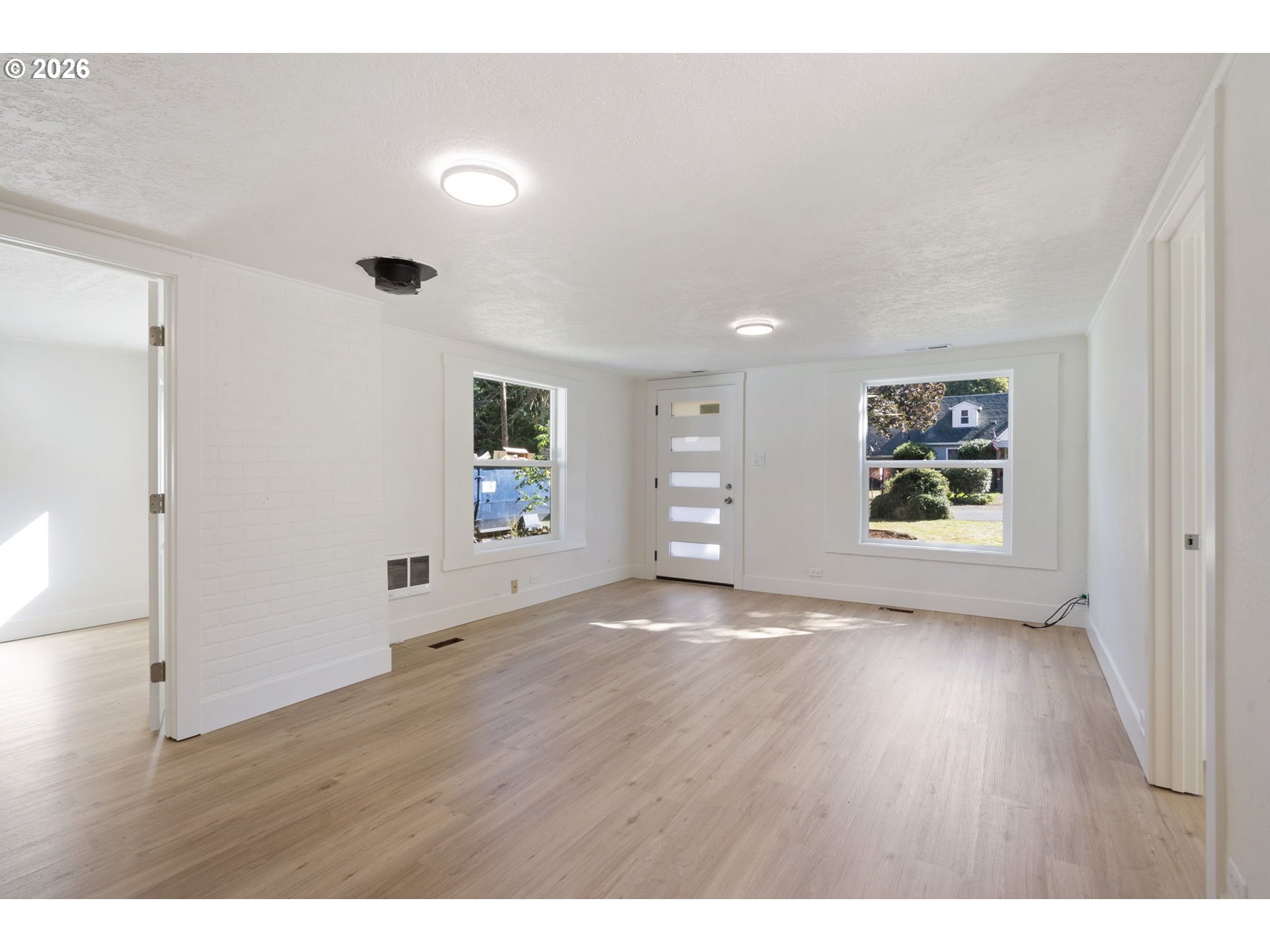 Vacant living room with light wood floors, white brick partition, and front door between two windows.