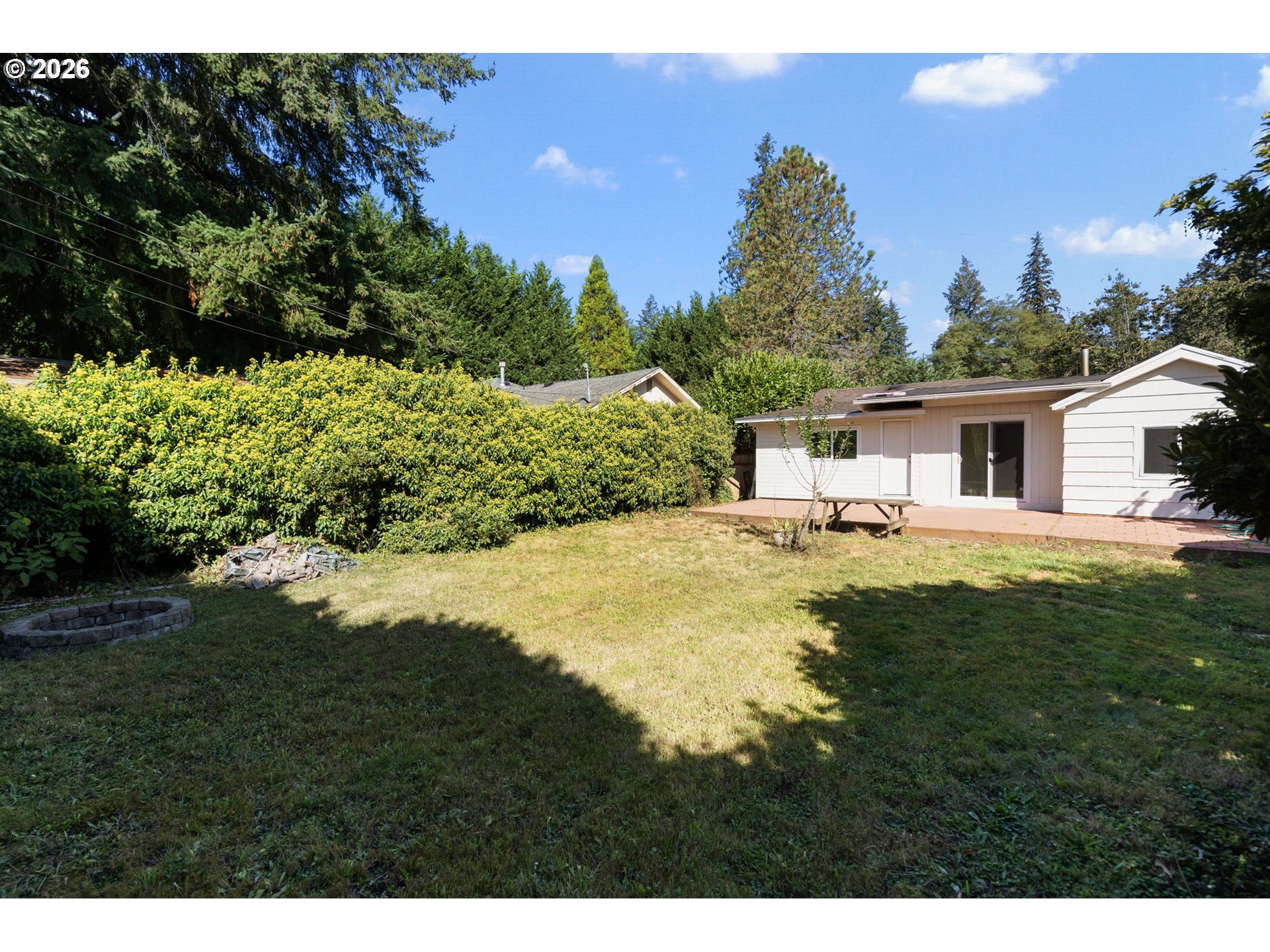 Rear of a white single-story house beyond a grassy backyard, with a patio and tall hedge.