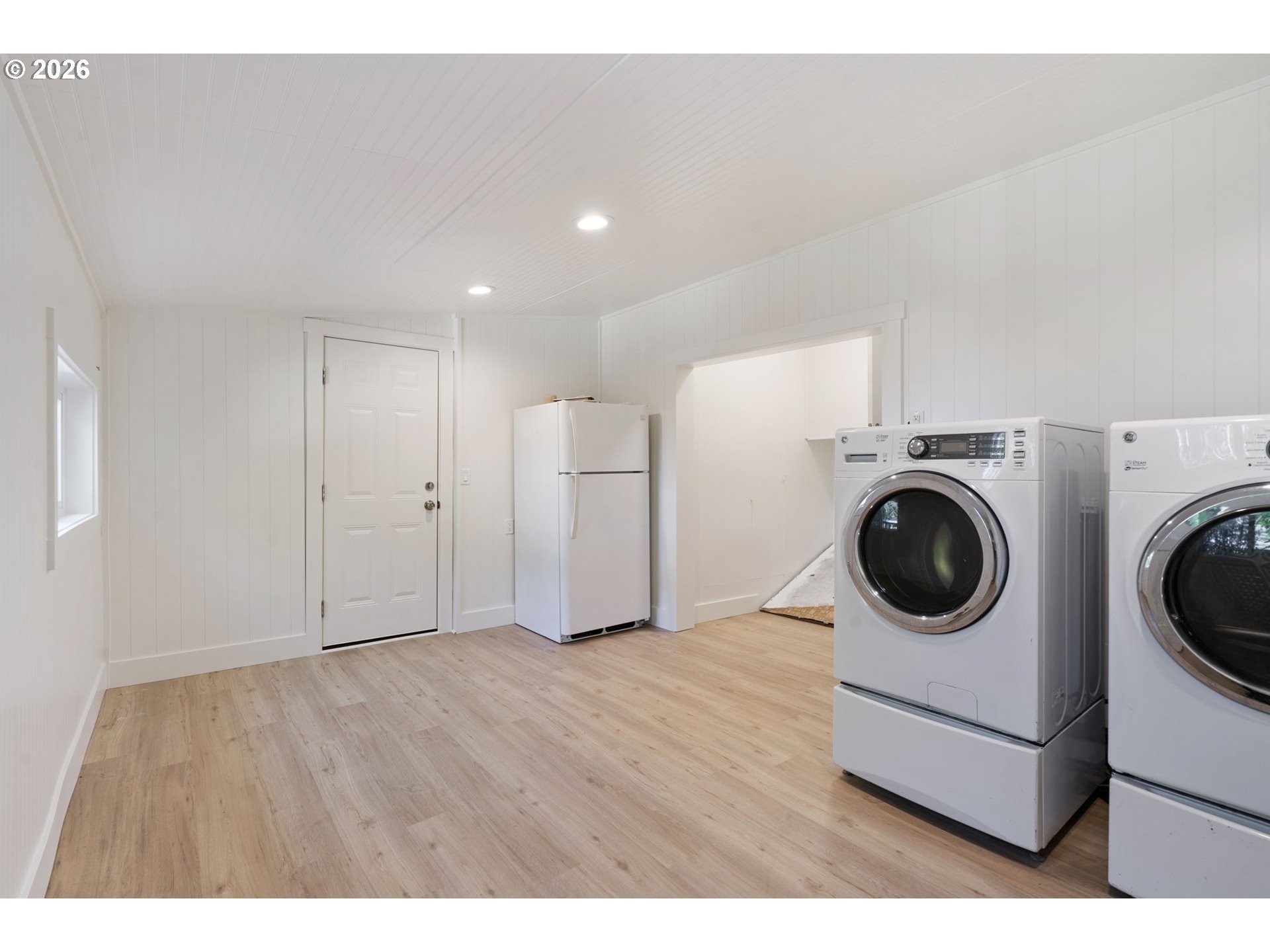 Laundry room with front-loading washer and dryer, refrigerator, and light wood flooring.