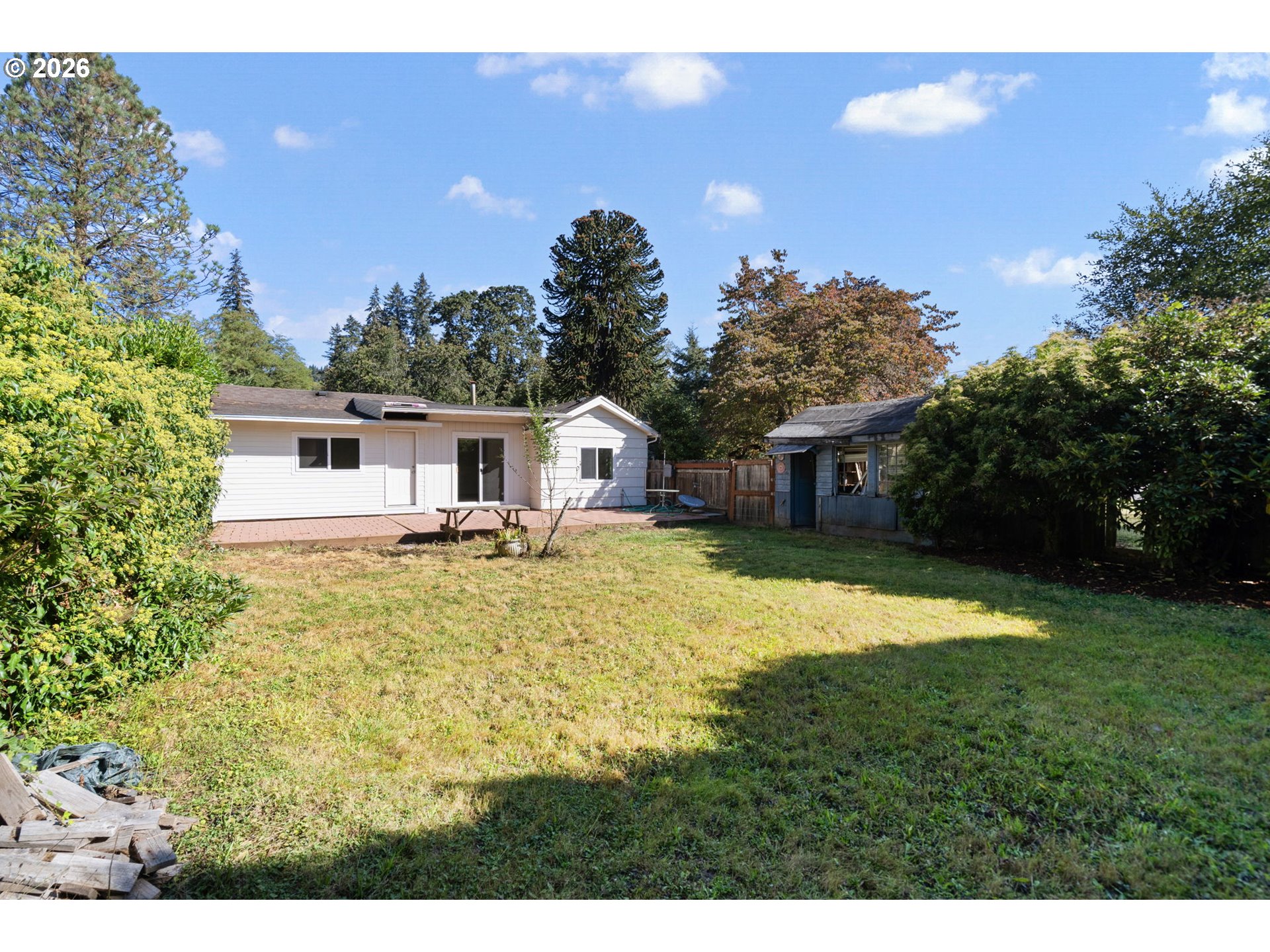 Backyard with a white house, deck, small outbuilding, and mature landscaping.
