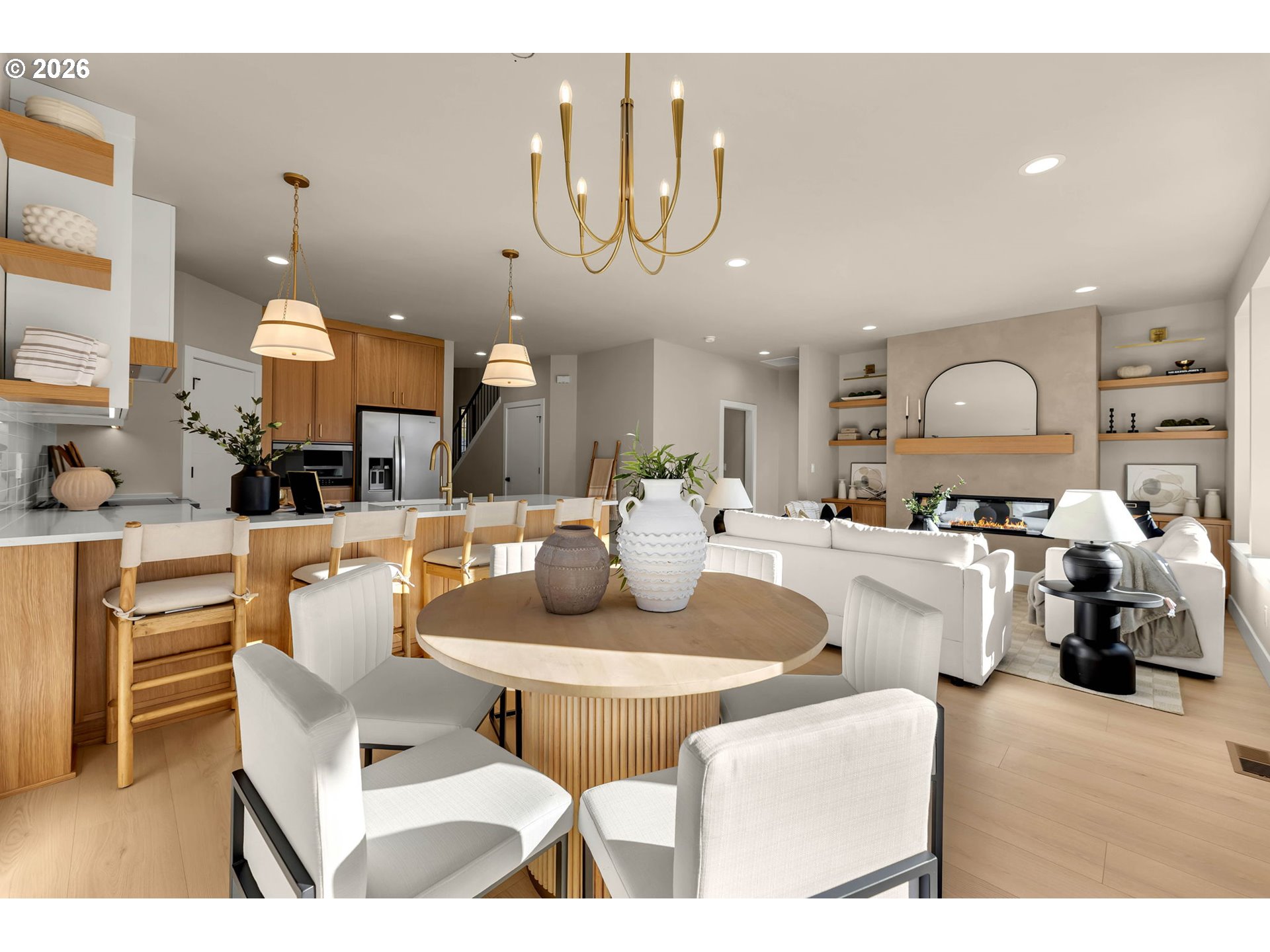 Open-plan kitchen, dining, and living room with light wood cabinetry, white seating, and a linear fireplace.