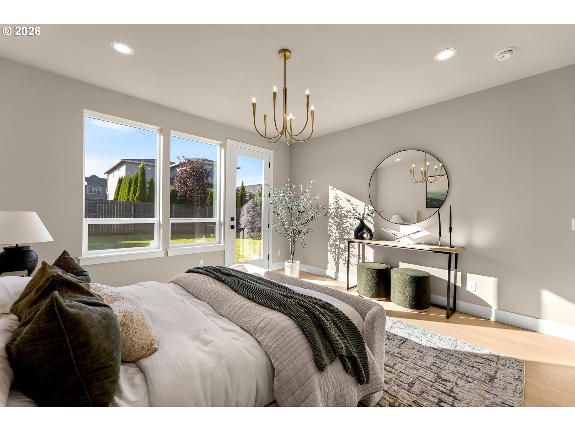 Staged bedroom with neutral bedding, large windows, a brass chandelier, and a round mirror.
