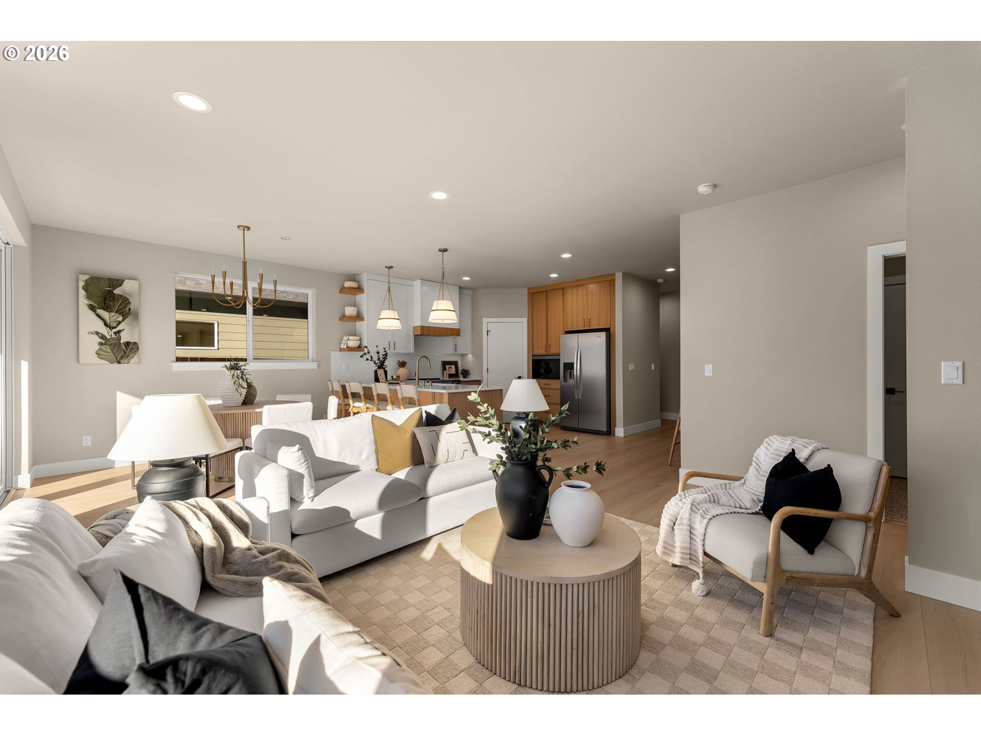 Open-plan living room and kitchen with white sofas, warm wood cabinets, and sunlight through large windows.