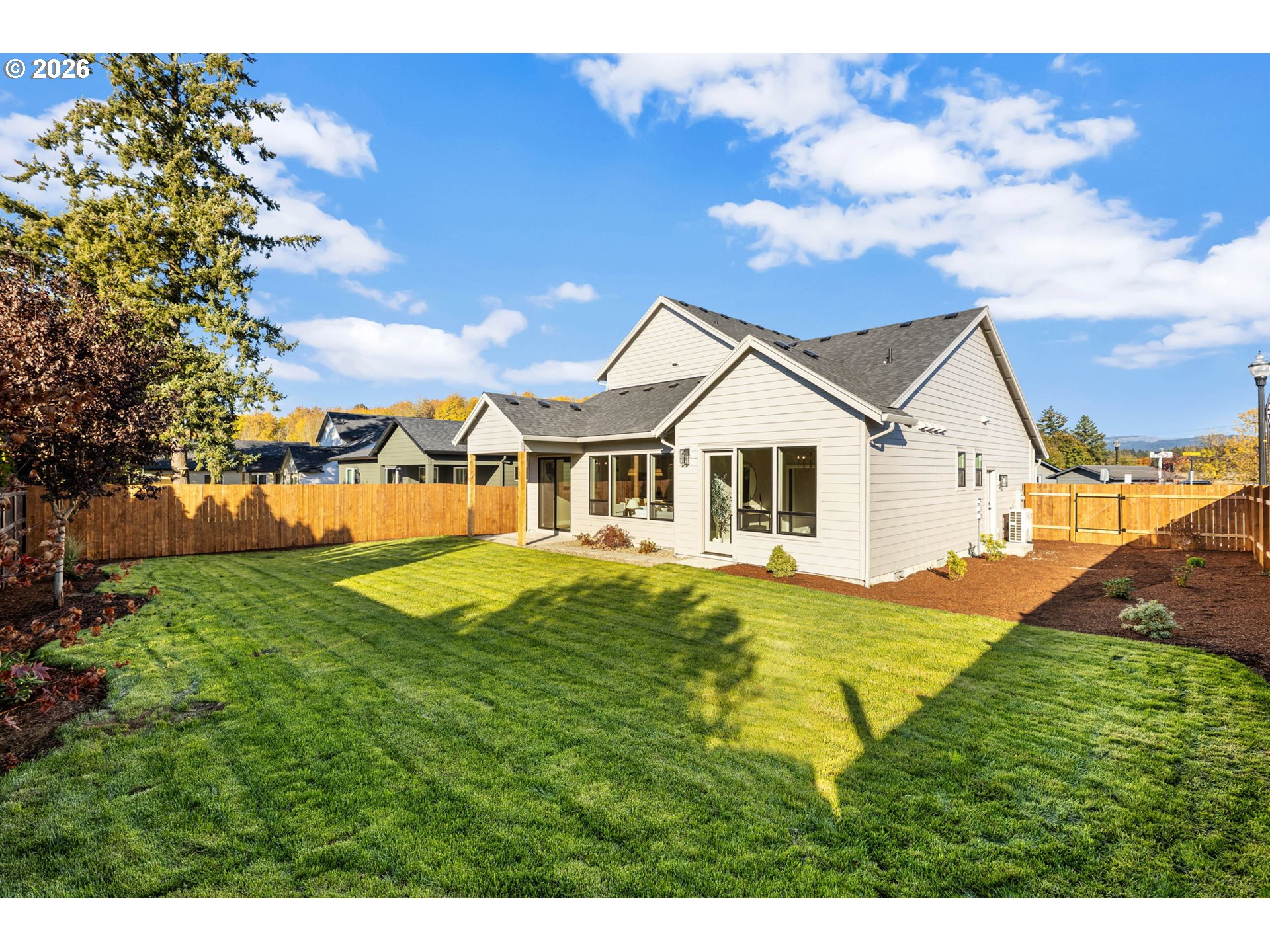 Rear of a light-colored house with a fenced backyard, broad lawn, and landscaped garden beds.