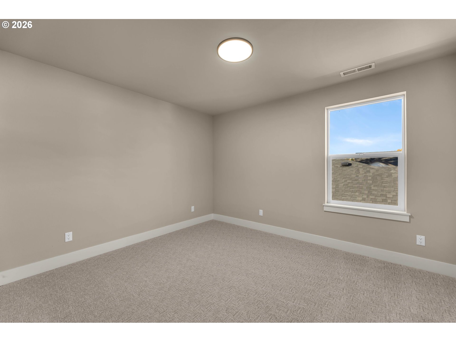 Empty bedroom with beige walls, patterned carpet, and a window overlooking a nearby roof.