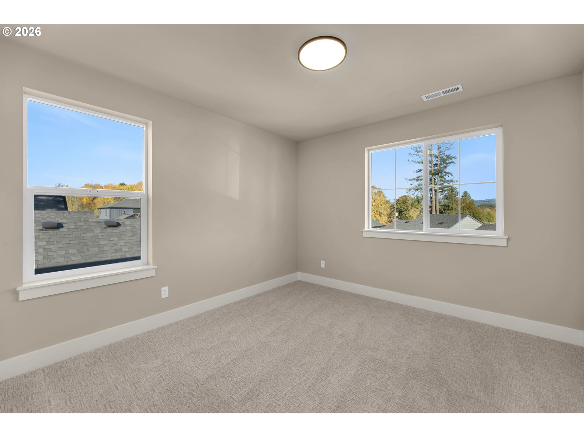 Empty carpeted bedroom with beige walls and two large windows overlooking autumn trees.