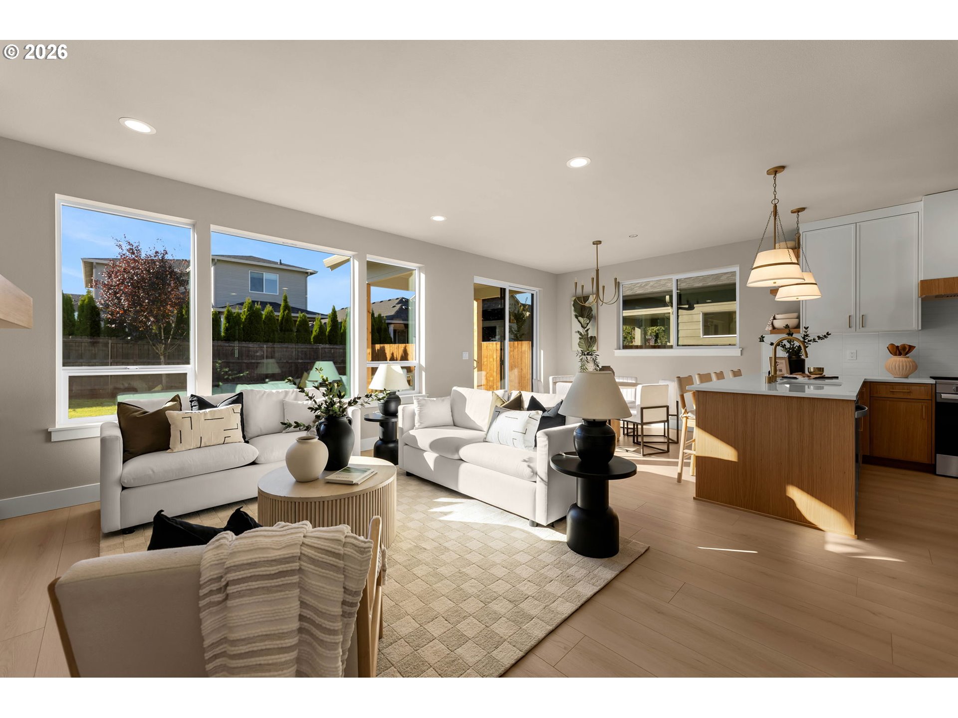 Open-plan living room with white sofas and a wood-front kitchen island beside large windows.