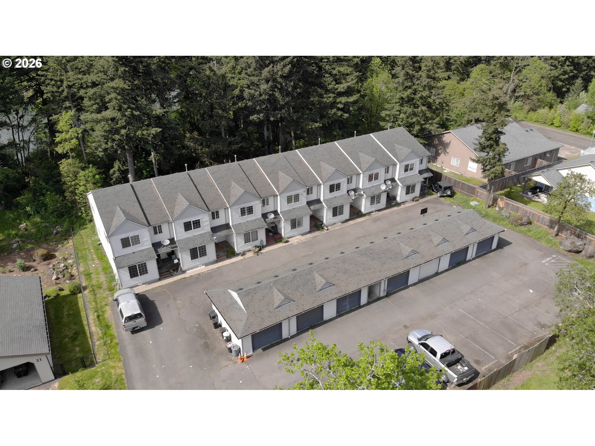 Aerial view of attached townhouses and a long garage building beside a wooded area.