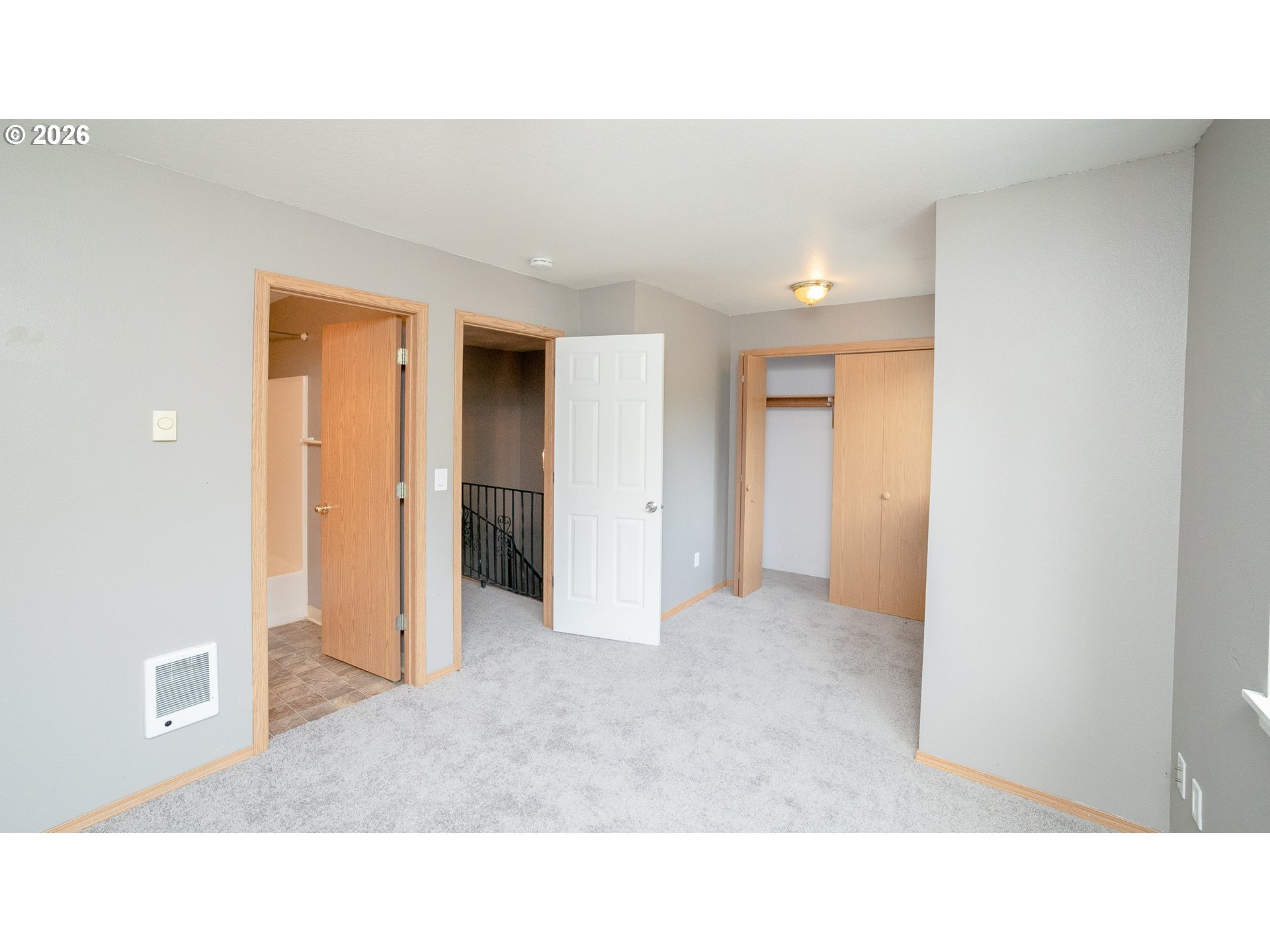 Vacant carpeted room with a closet, tiled bathroom doorway, and stair landing with black railing.