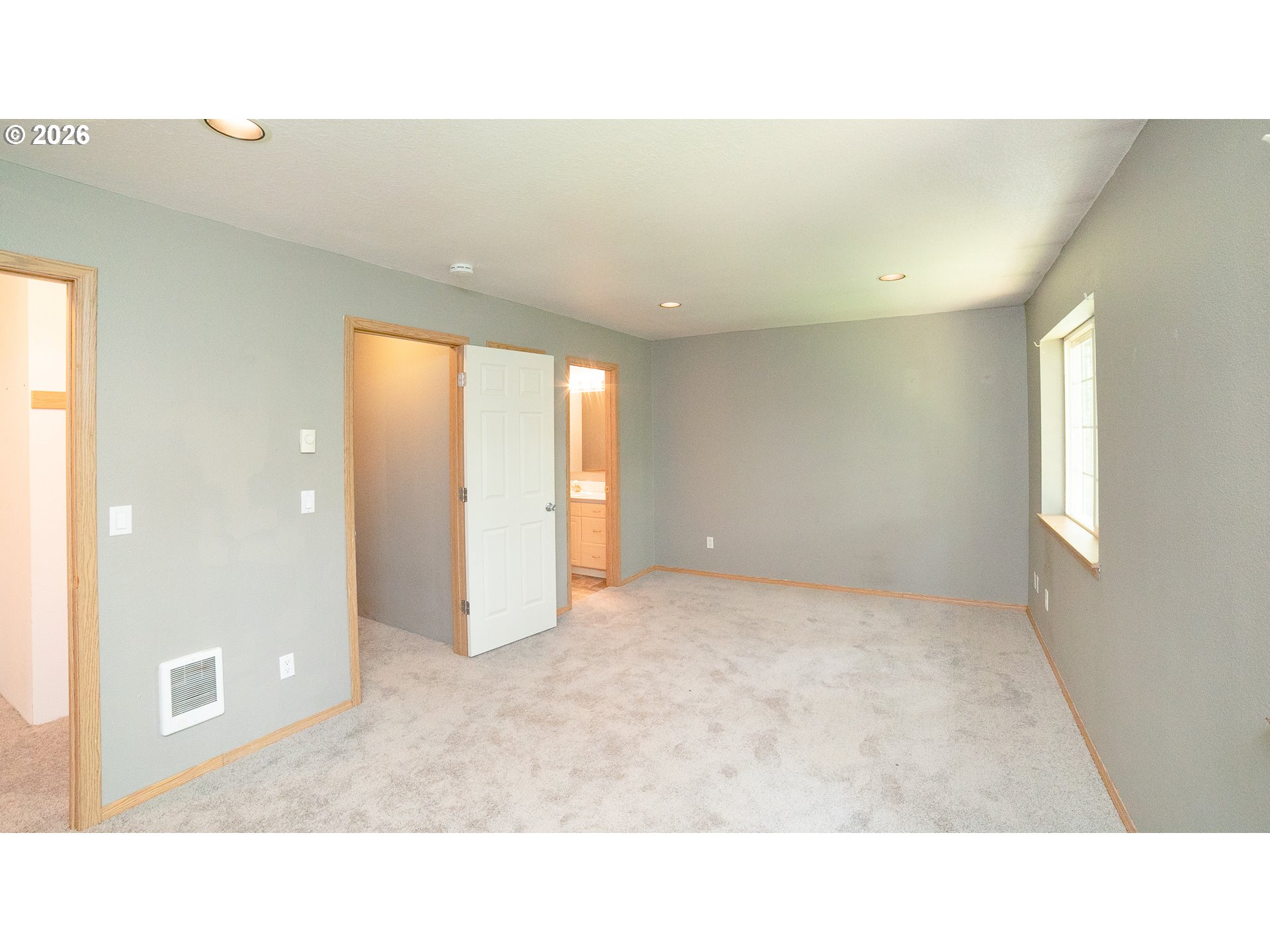 Empty carpeted bedroom with gray walls, a window, and an open bathroom doorway.