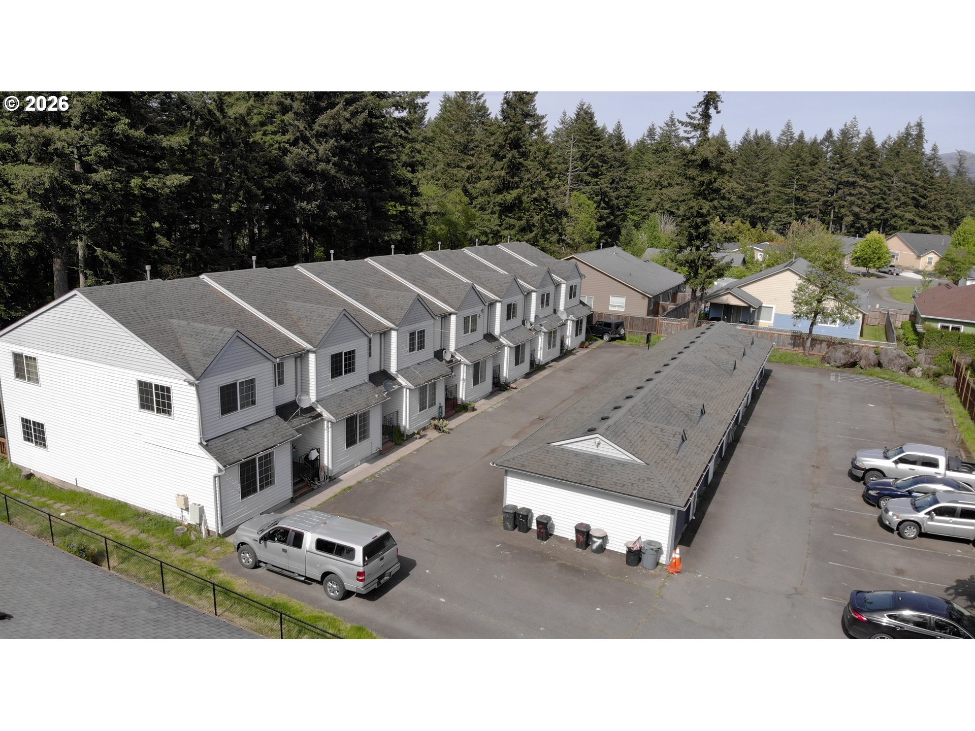 Aerial view of multi-unit homes and parking area bordered by evergreen trees.