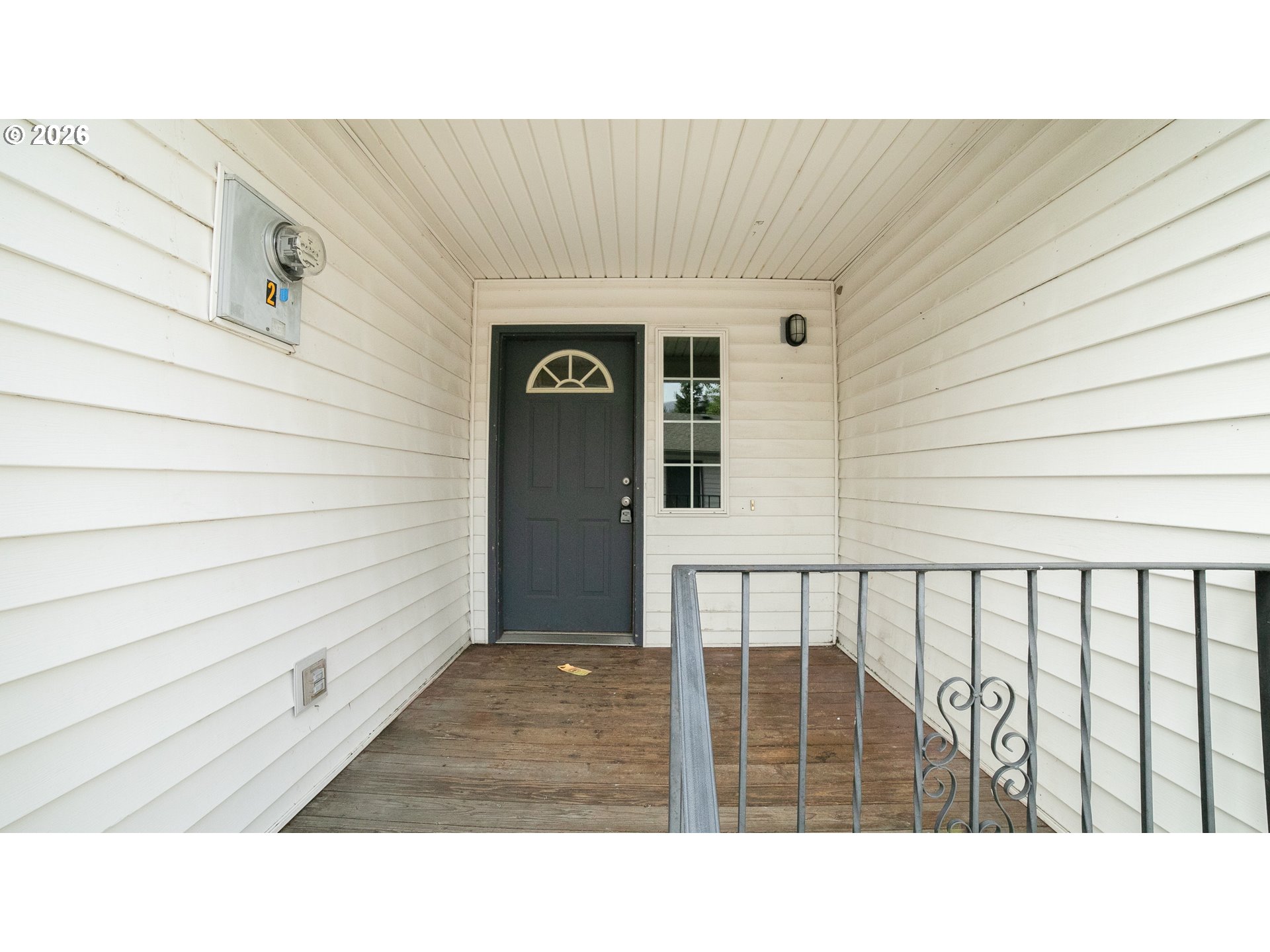 Covered front entry with a dark door, white siding, wood decking, and metal railing.