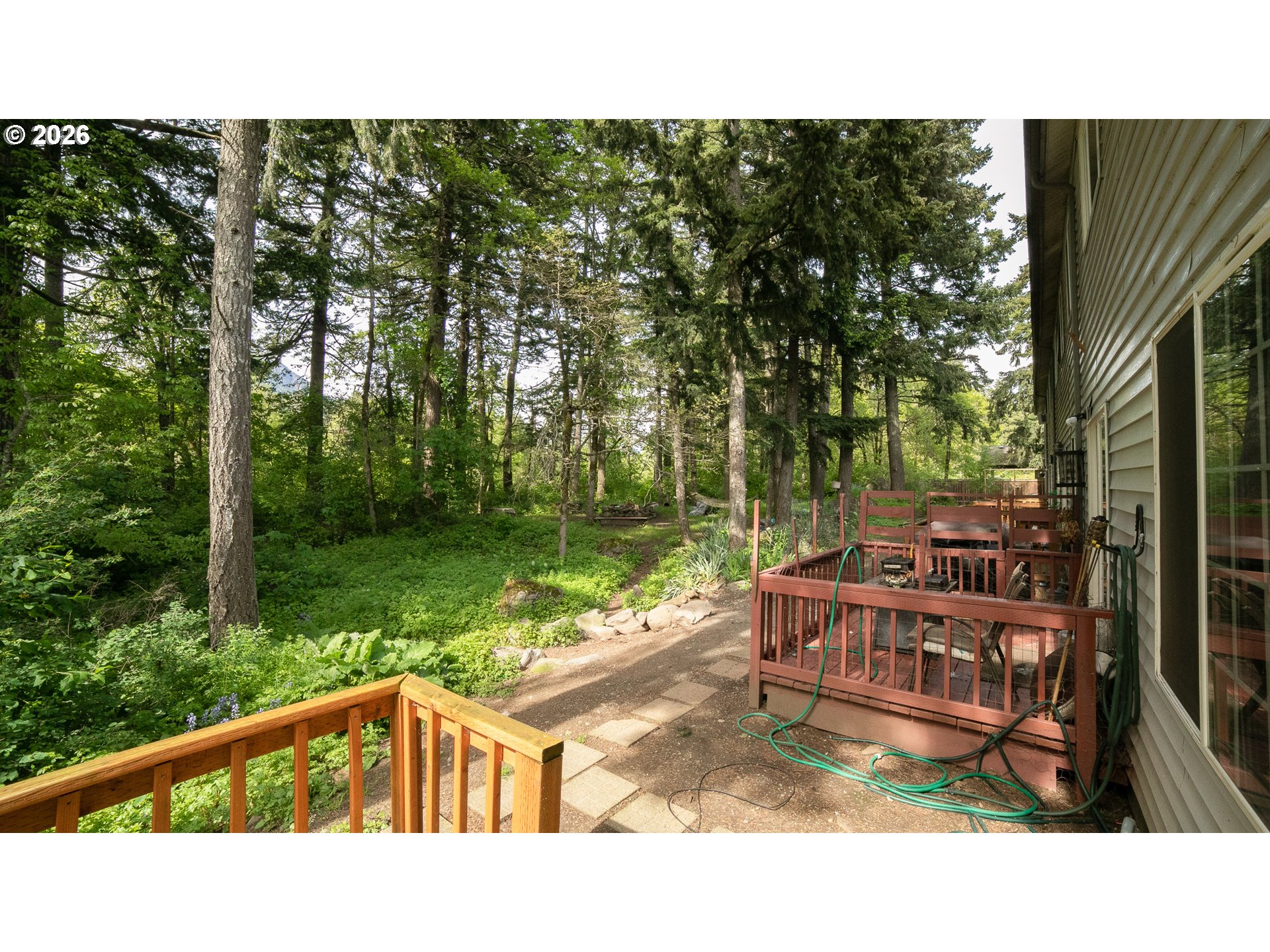 Wooded backyard with a raised deck, outdoor seating, and a paved garden path.