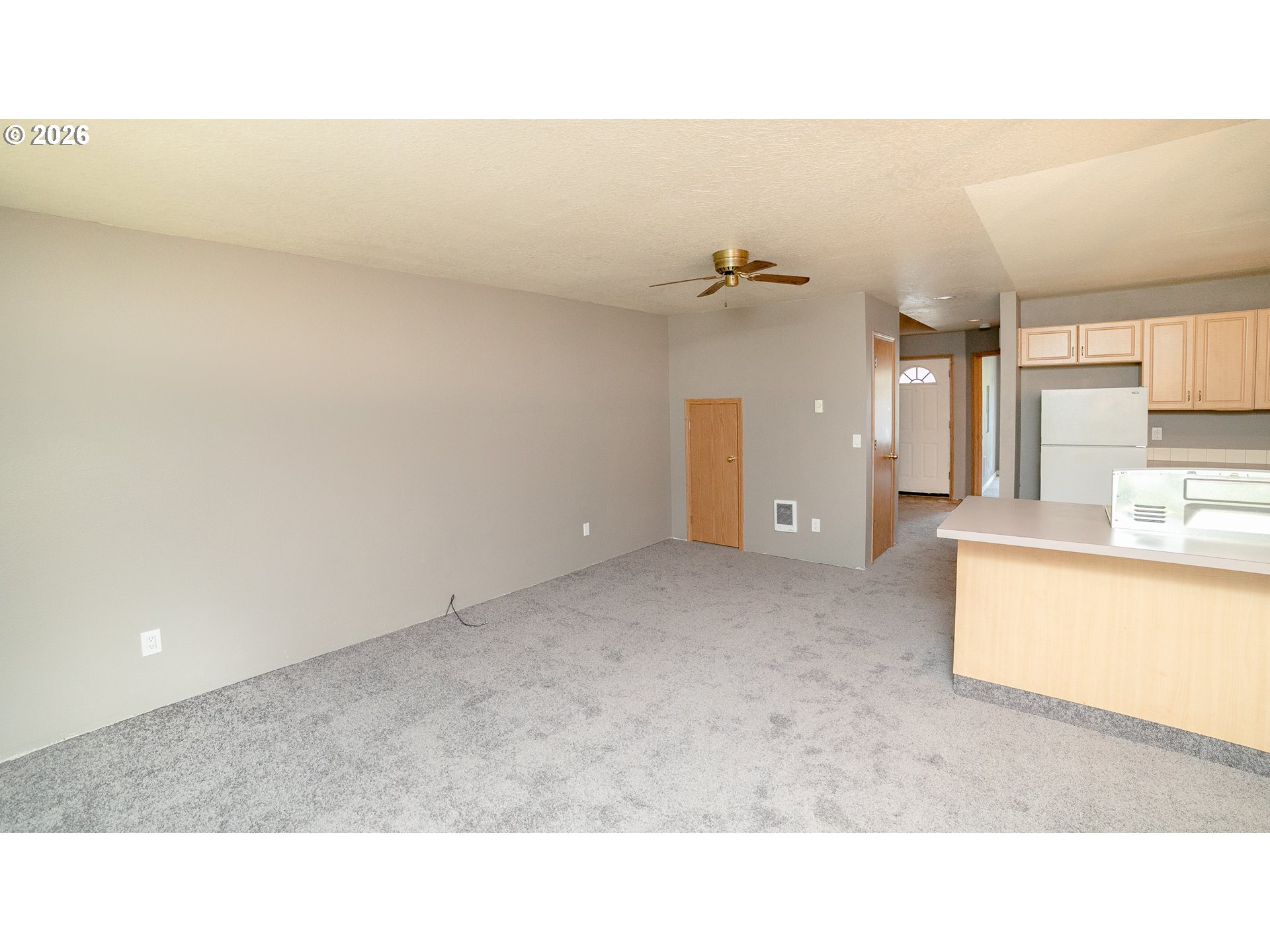 Unfurnished living area with gray carpet, ceiling fan, and open kitchen with wood cabinets and breakfast counter.