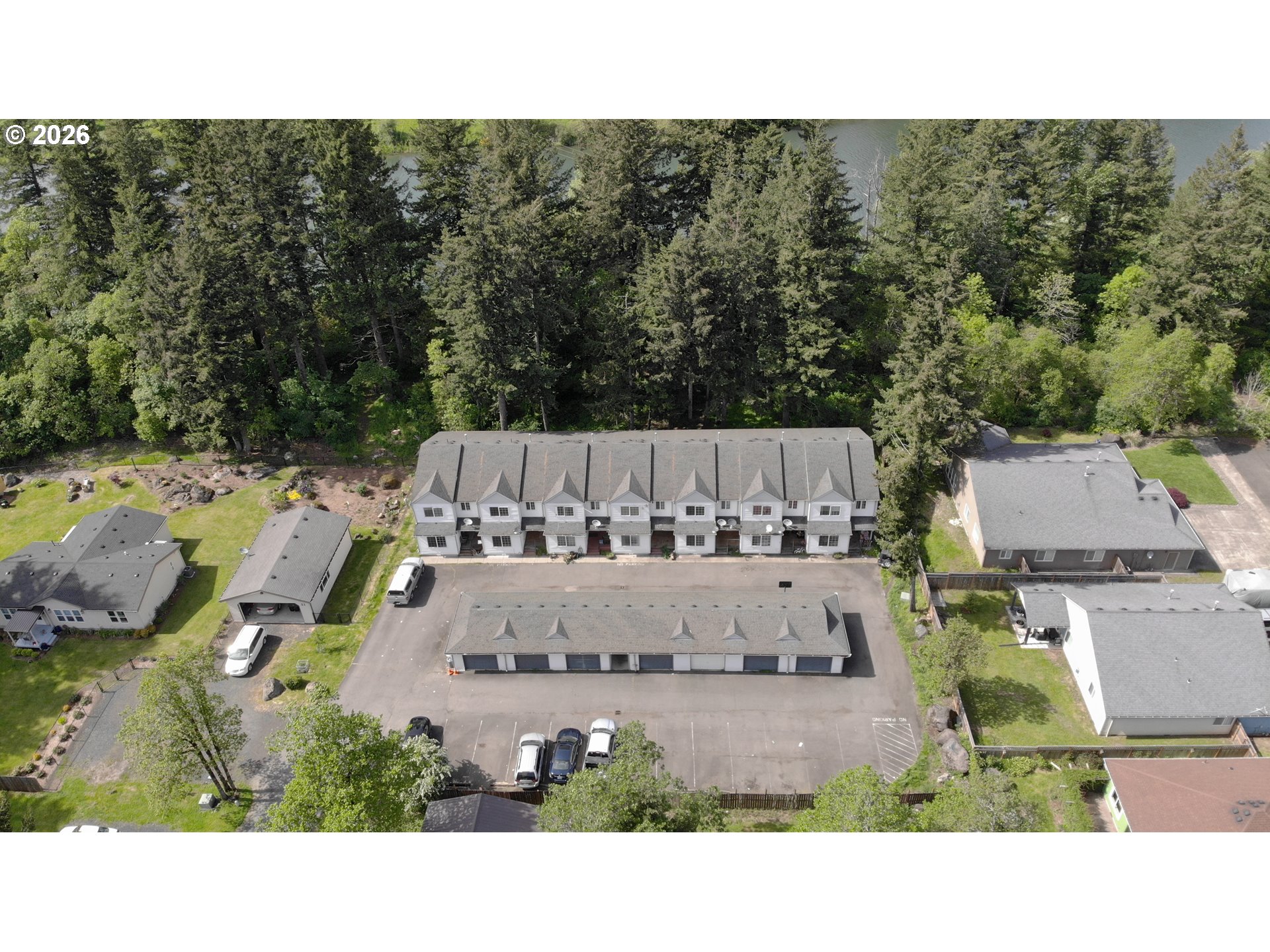 Aerial view of attached homes and parking lot bordered by dense trees, with water beyond.
