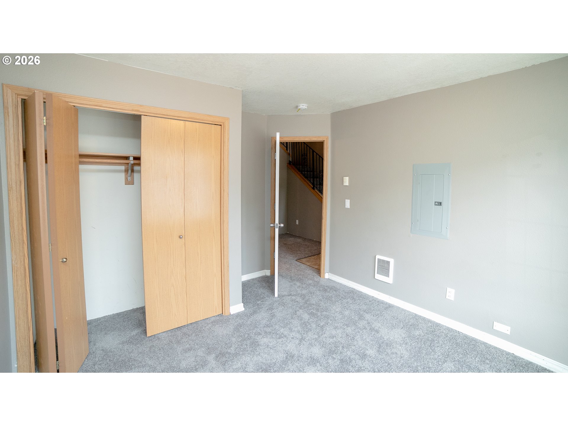 Empty carpeted bedroom with an open wooden closet and a staircase visible through the doorway.