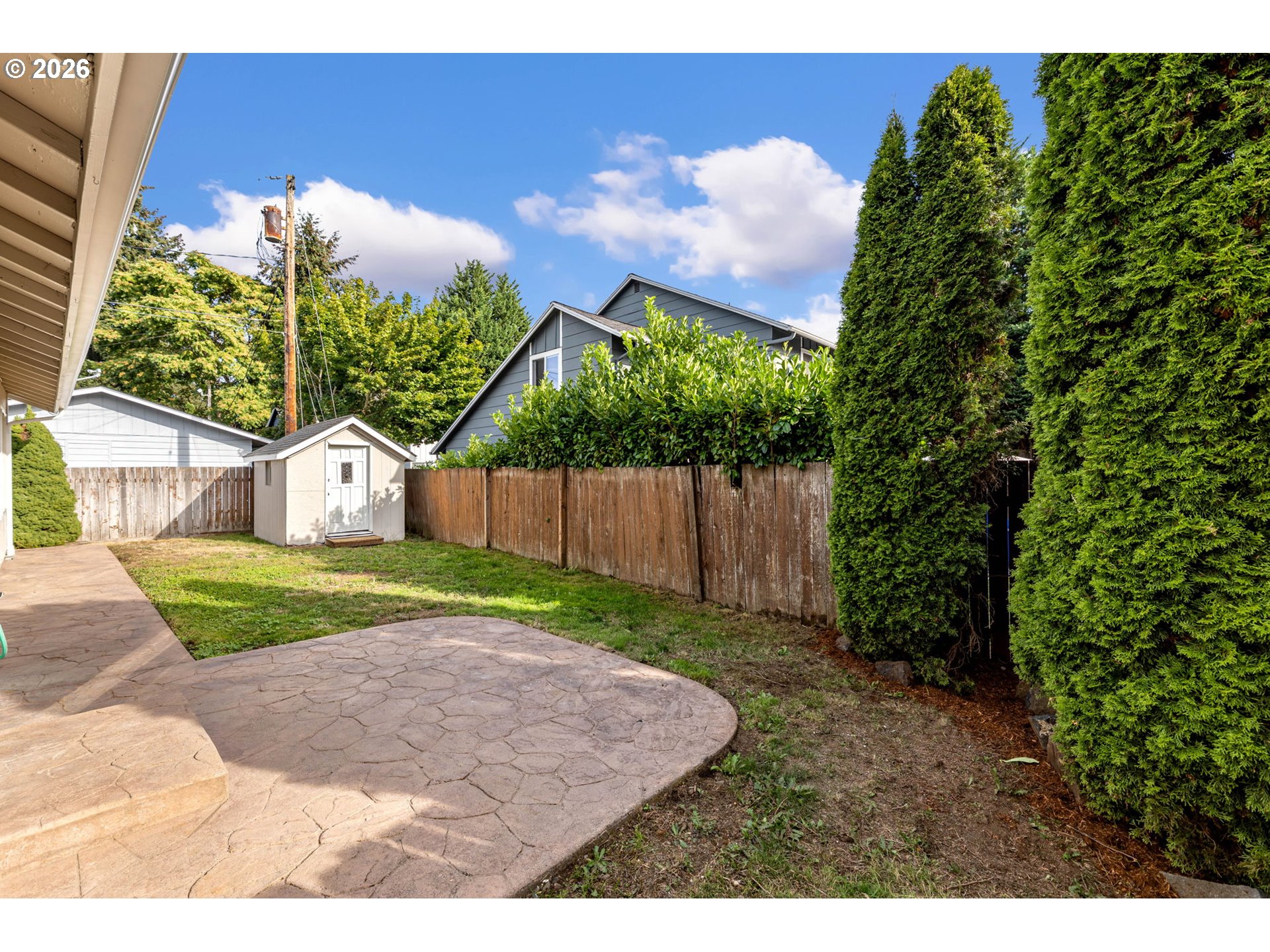Backyard with a stamped-concrete patio, small shed, and tall evergreen hedges.