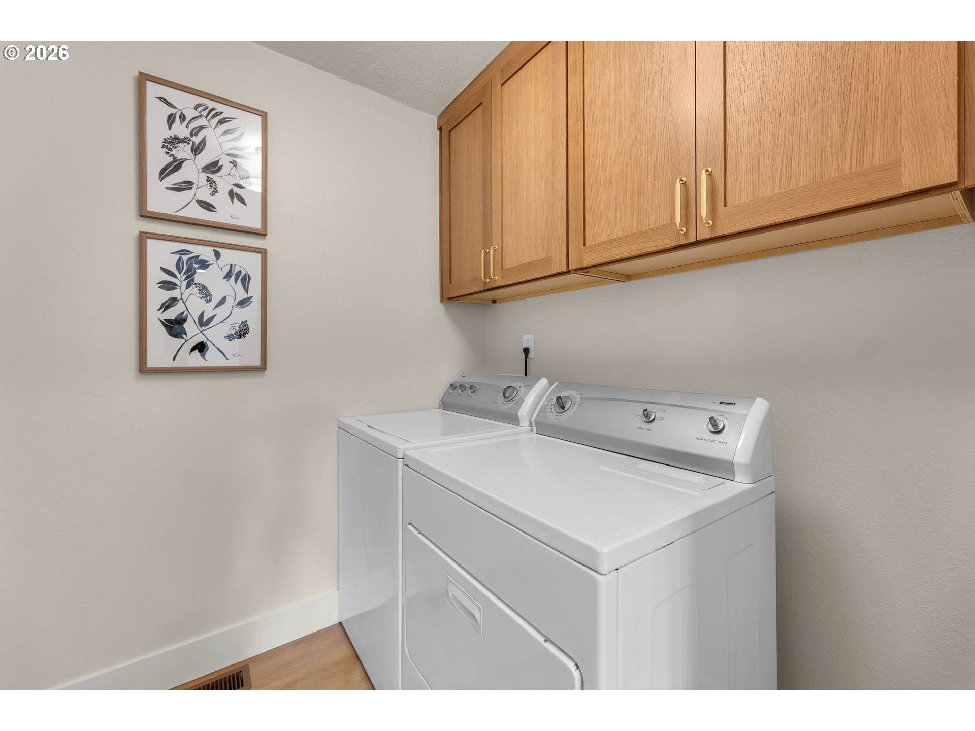 Laundry room with white washer and dryer beneath oak cabinets and framed botanical prints.
