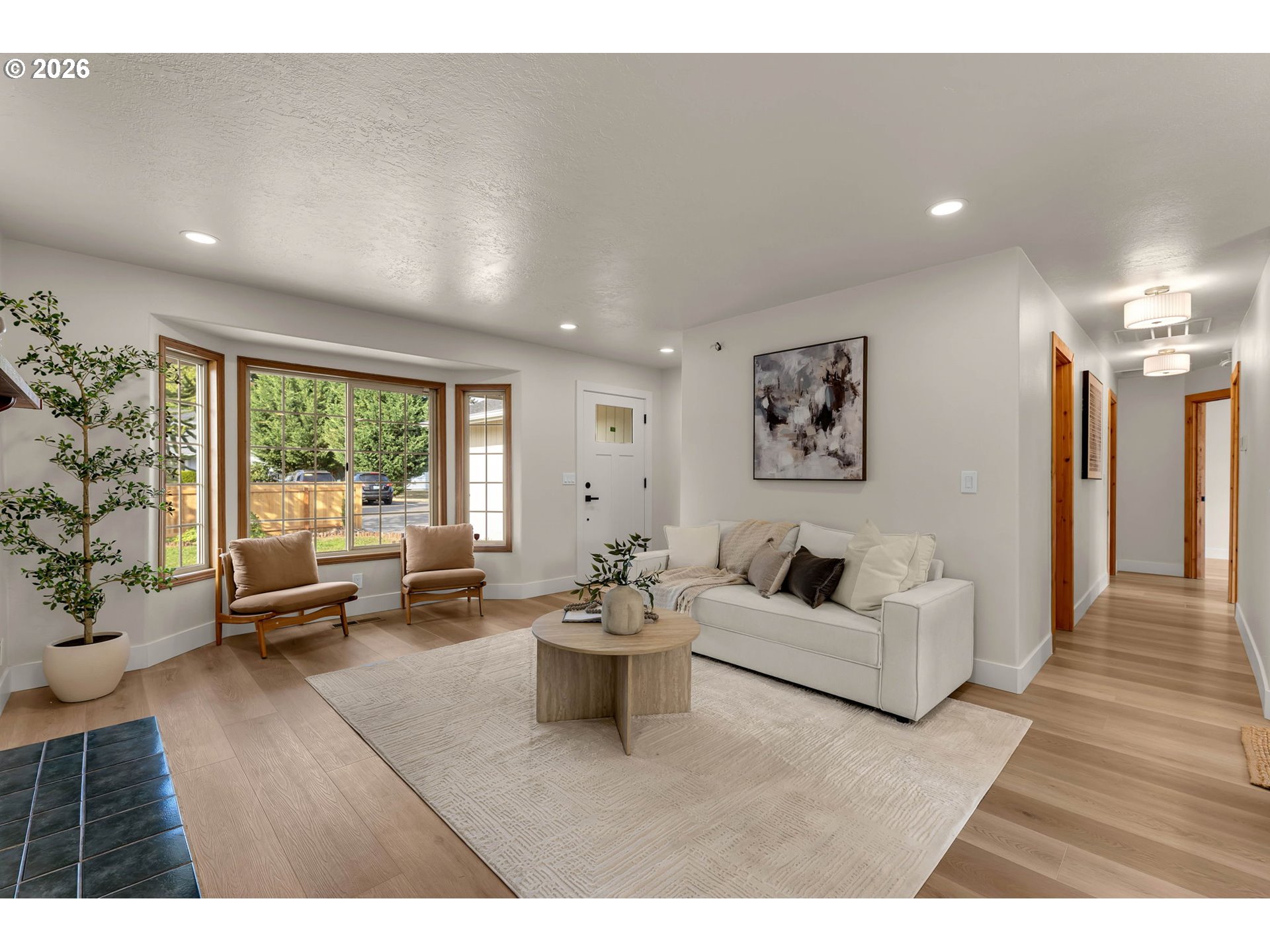 Staged living room with a white sofa, wood-framed chairs, and large windows overlooking greenery.