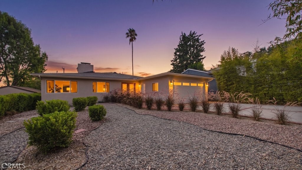 Single-story home at dusk with glowing windows, a gravel yard, and ornamental grasses.