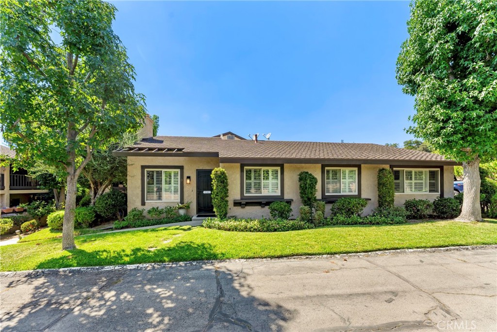 Single-story stucco house with dark trim, shuttered windows, and mature trees framing its front lawn.