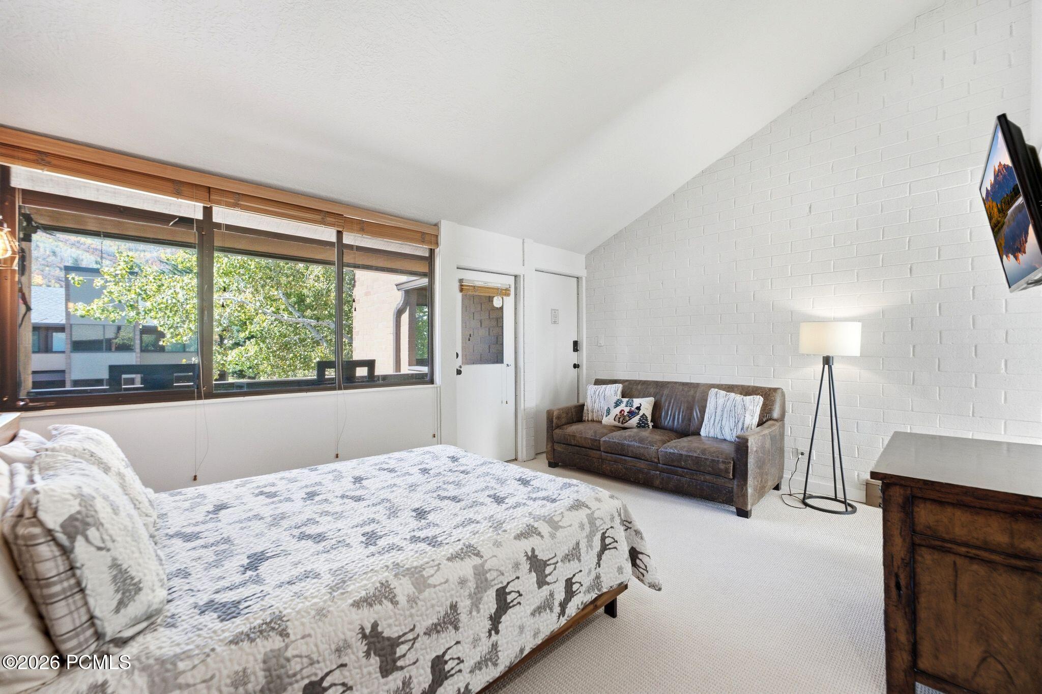Bedroom with a patterned bedspread, brown loveseat, and large windows overlooking trees.