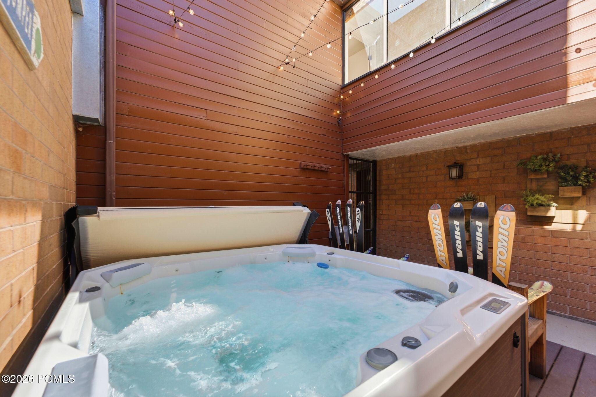 Outdoor hot tub on a brick patio, with skis stored nearby and string lights overhead.