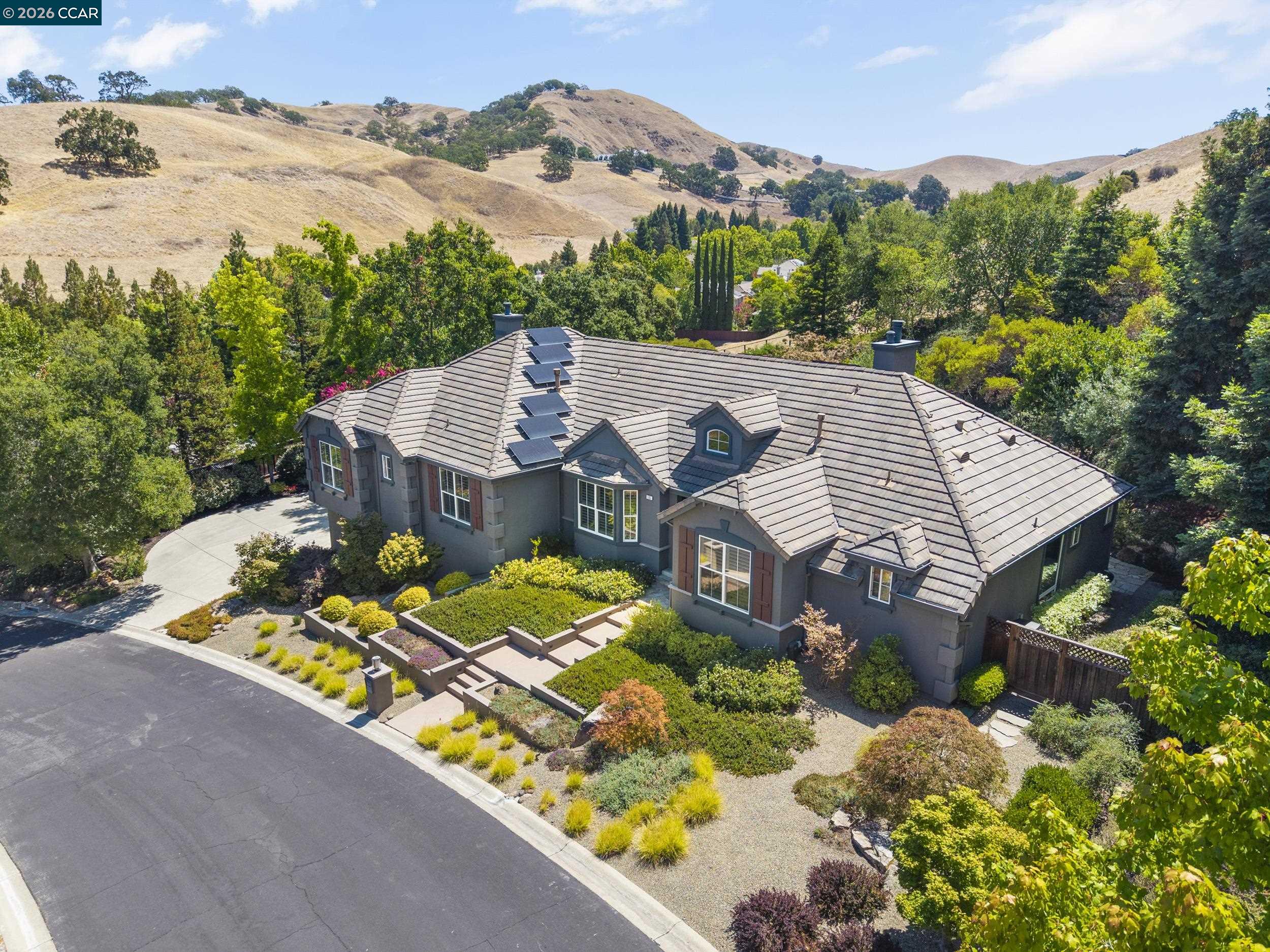 Aerial view of a gray home with roof solar panels, landscaped gardens, and rolling hills behind it.