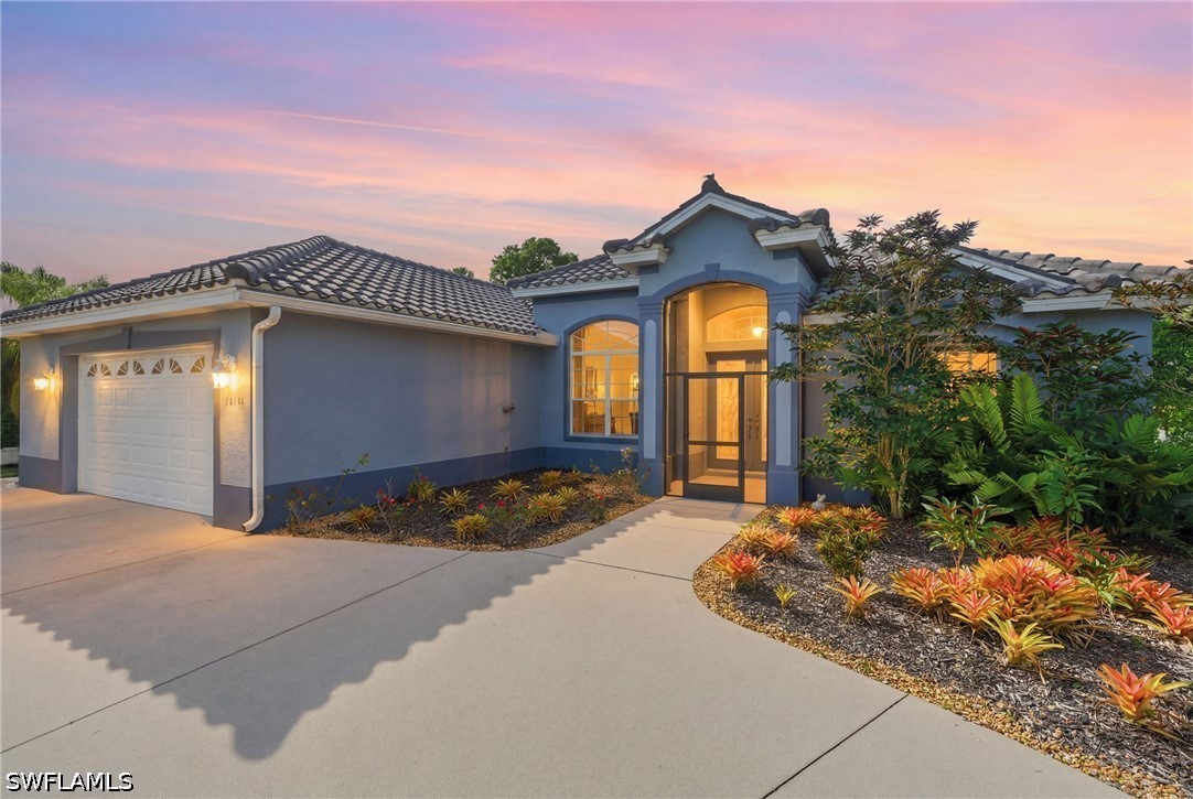 Blue-gray single-story home with an illuminated entry, tiled roof, and colorful front landscaping at dusk.