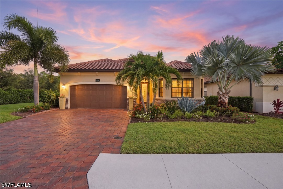 Single-story home with a brick paver driveway, tropical landscaping, and a pink dusk sky.