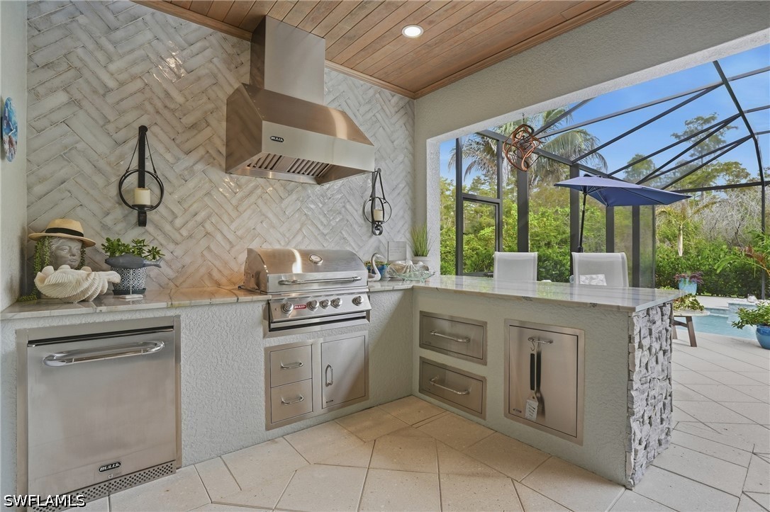 Outdoor kitchen with stainless steel appliances and stone backsplash beside a screened pool lanai.