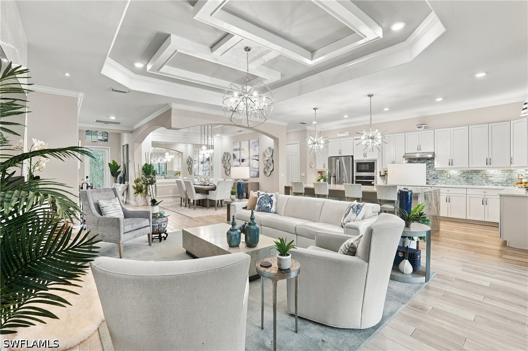 Open-plan living room with pale furnishings, a geometric tray ceiling, and adjoining white kitchen.