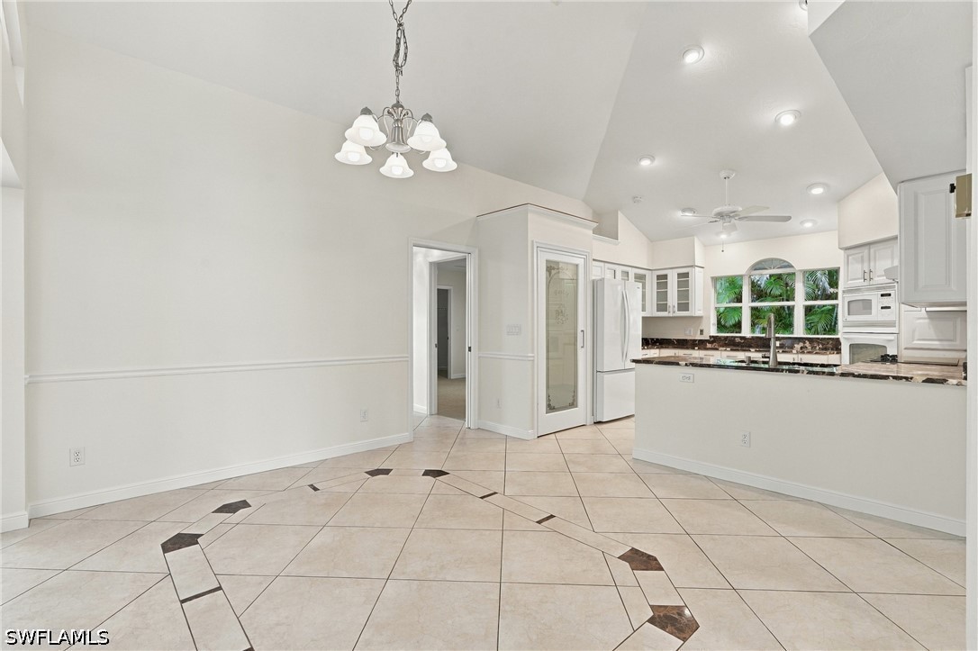 Empty tiled dining area opening into a white kitchen with dark countertops and tropical greenery beyond the windows.