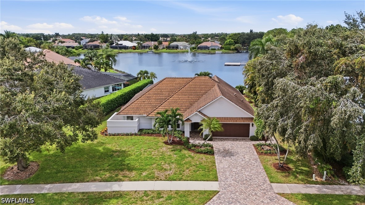 Aerial view of a tile-roofed home beside a lake with a fountain and paver driveway.