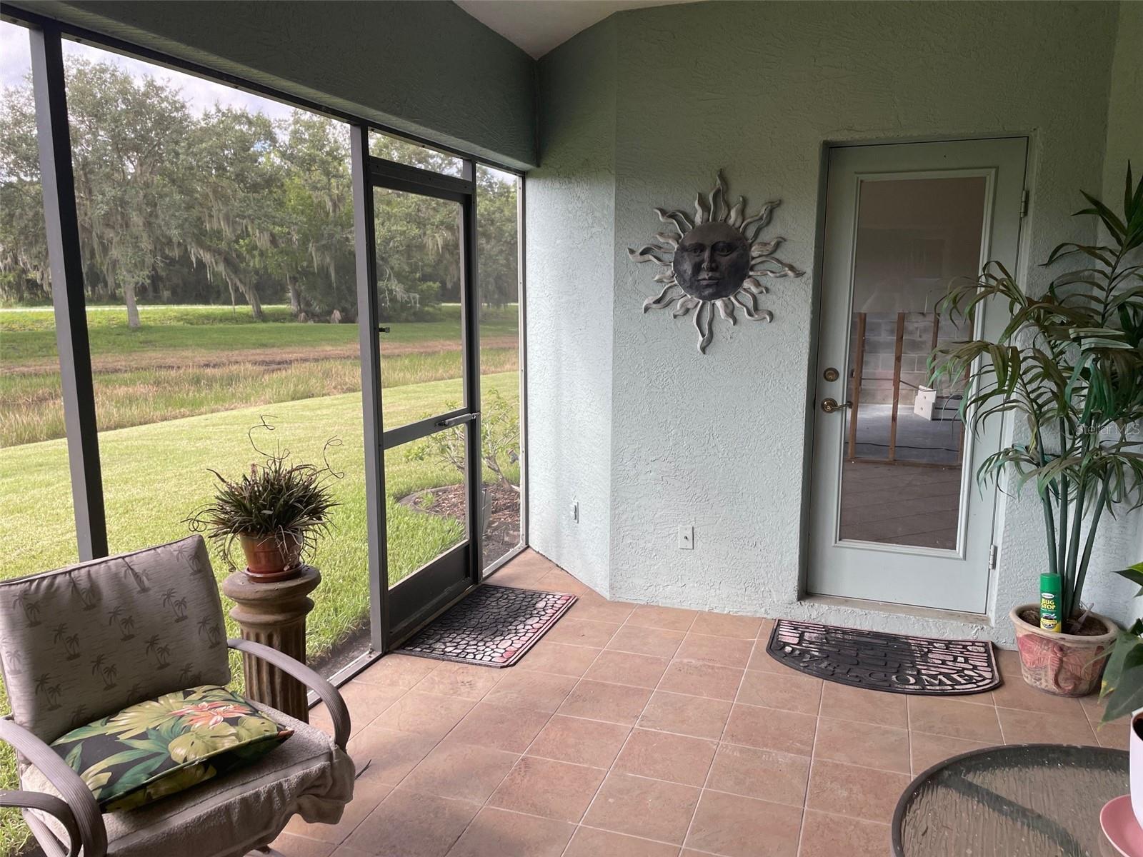 Screened lanai with tiled floor, patio seating, and views of a grassy yard and trees.