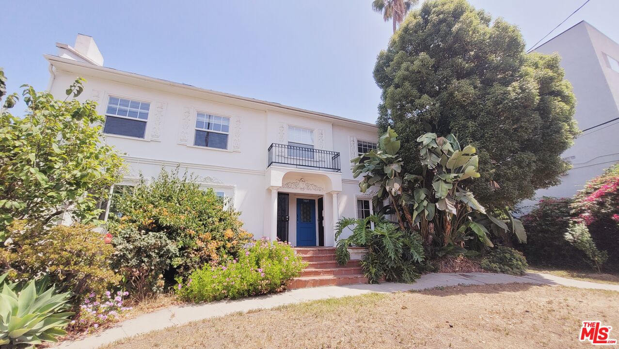 White two-story house with a blue front door, small balcony, and lush landscaping.