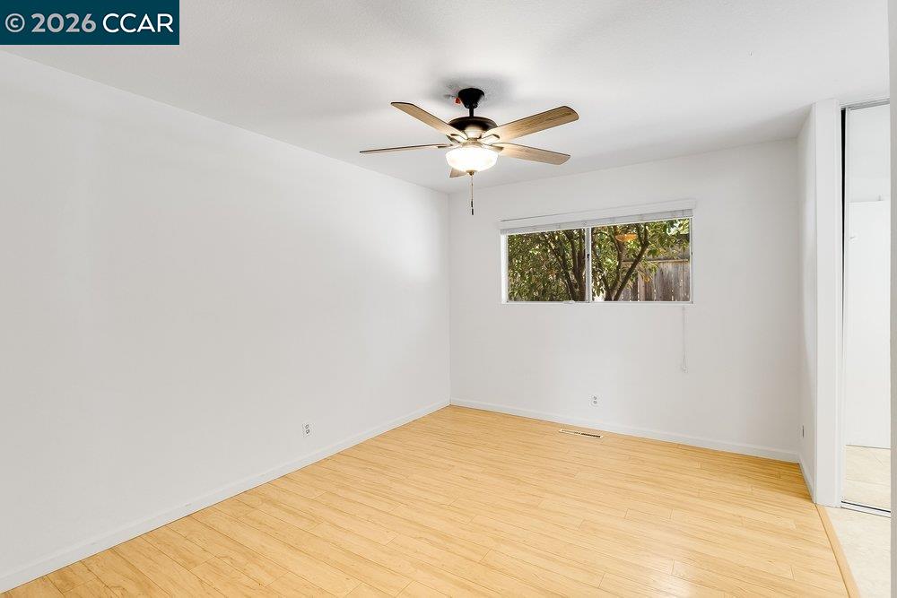 Empty room with light wood flooring, a ceiling fan, and a window overlooking trees.